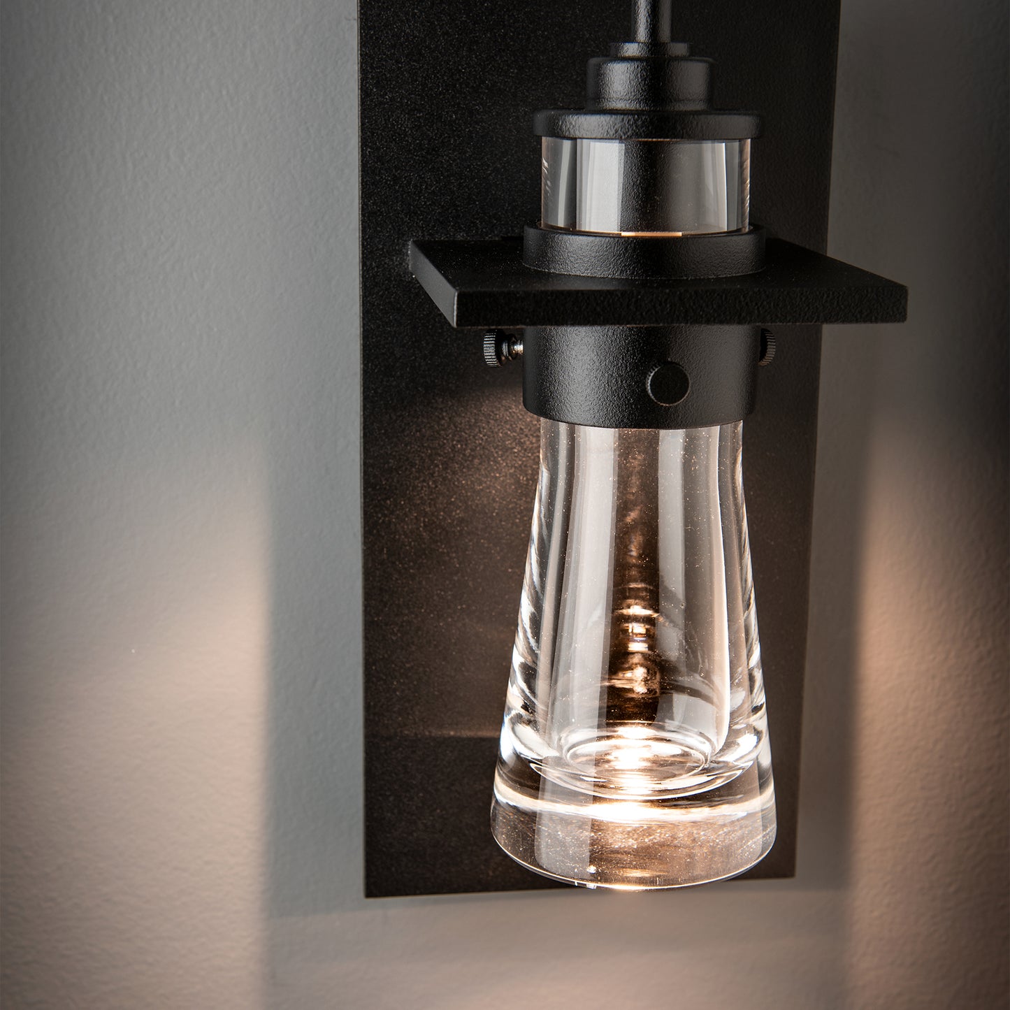 The Erlenmeyer Medium Outdoor Sconce by Hubbardton Forge showcases a modern cylindrical glass shade and sturdy black metal craftsmanship.