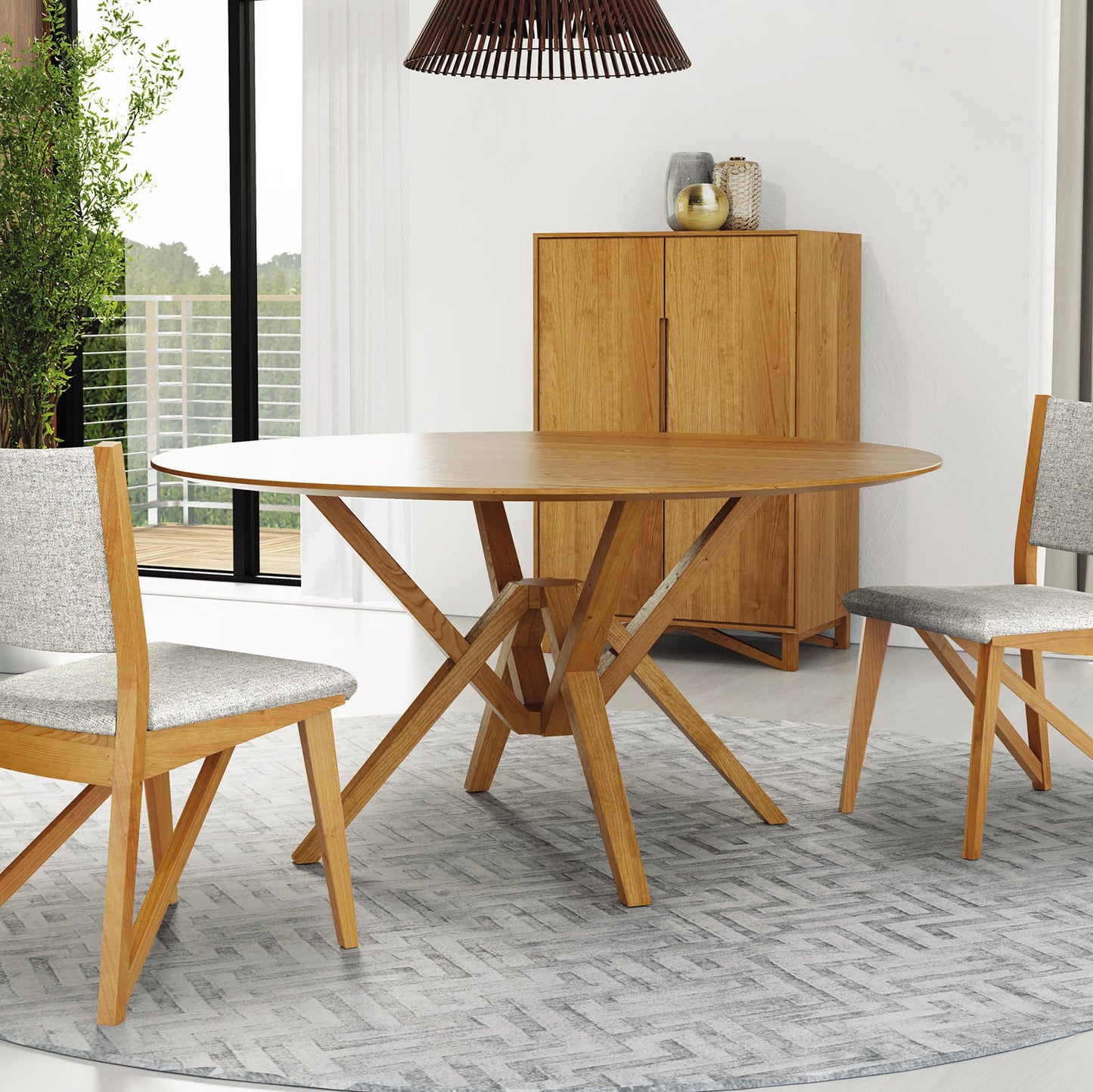 A modern dining room features a round wooden table with an intricate base, two matching chairs with light gray cushions, a light gray rug with geometric patterns, the Exeter Bar Cabinet by Copeland Furniture, a potted plant, large windows with a view of a balcony, and a hanging light fixture.