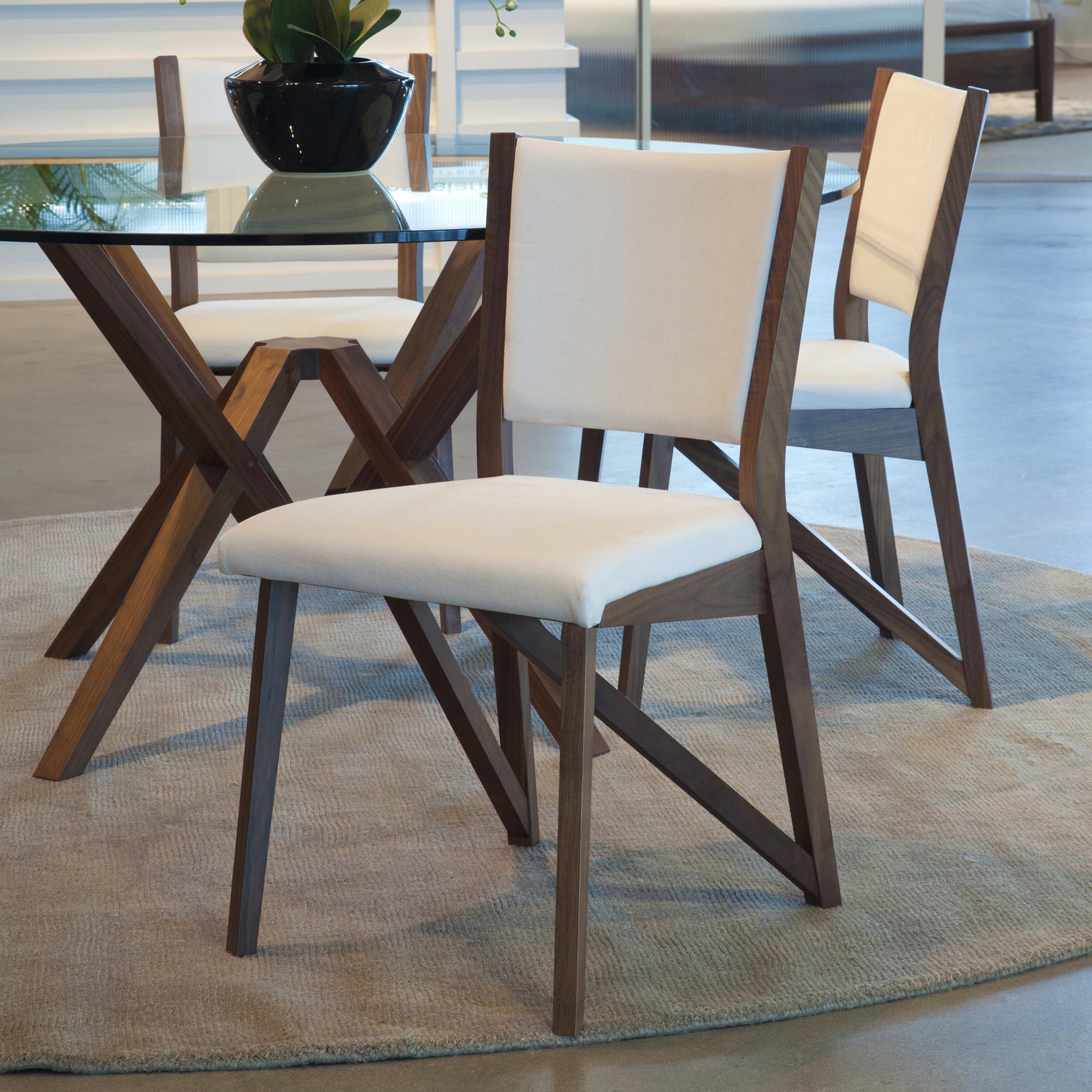 Two Copeland Furniture Exeter Chairs with ergonomic seat backs and white cushions are placed around a glass-top table with a wooden base. The table is set on a beige rug in a contemporary room with a polished concrete floor. A plant in a black pot sits on the table.