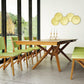 A modern dining room showcases the Exeter Extension Dining Table from Copeland Furniture, crafted from sustainably harvested solid North American hardwood. The table, with its unique geometric base, is surrounded by green upholstered chairs. A large window offers a view of trees, complemented by gold circular wall discs and candle holders on the table.