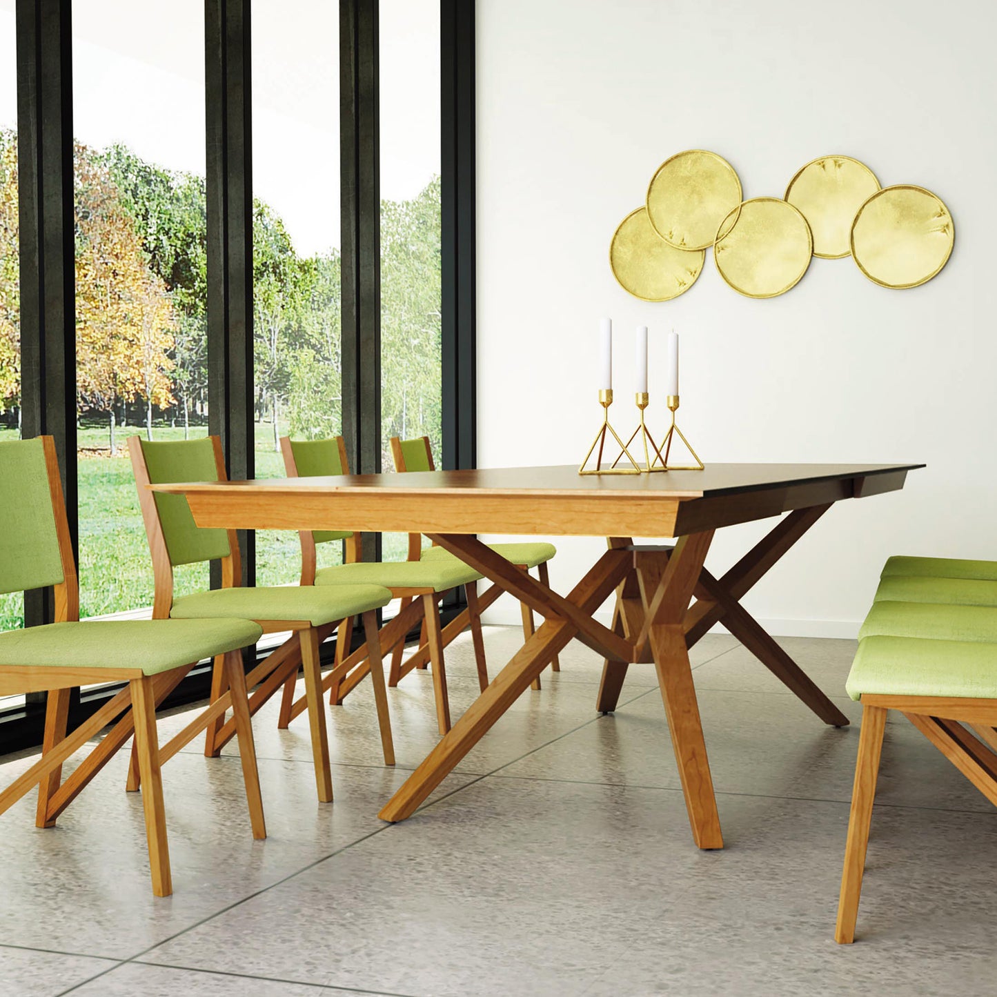 A modern dining room showcases the Exeter Extension Dining Table from Copeland Furniture, crafted from sustainably harvested solid North American hardwood. The table, with its unique geometric base, is surrounded by green upholstered chairs. A large window offers a view of trees, complemented by gold circular wall discs and candle holders on the table.