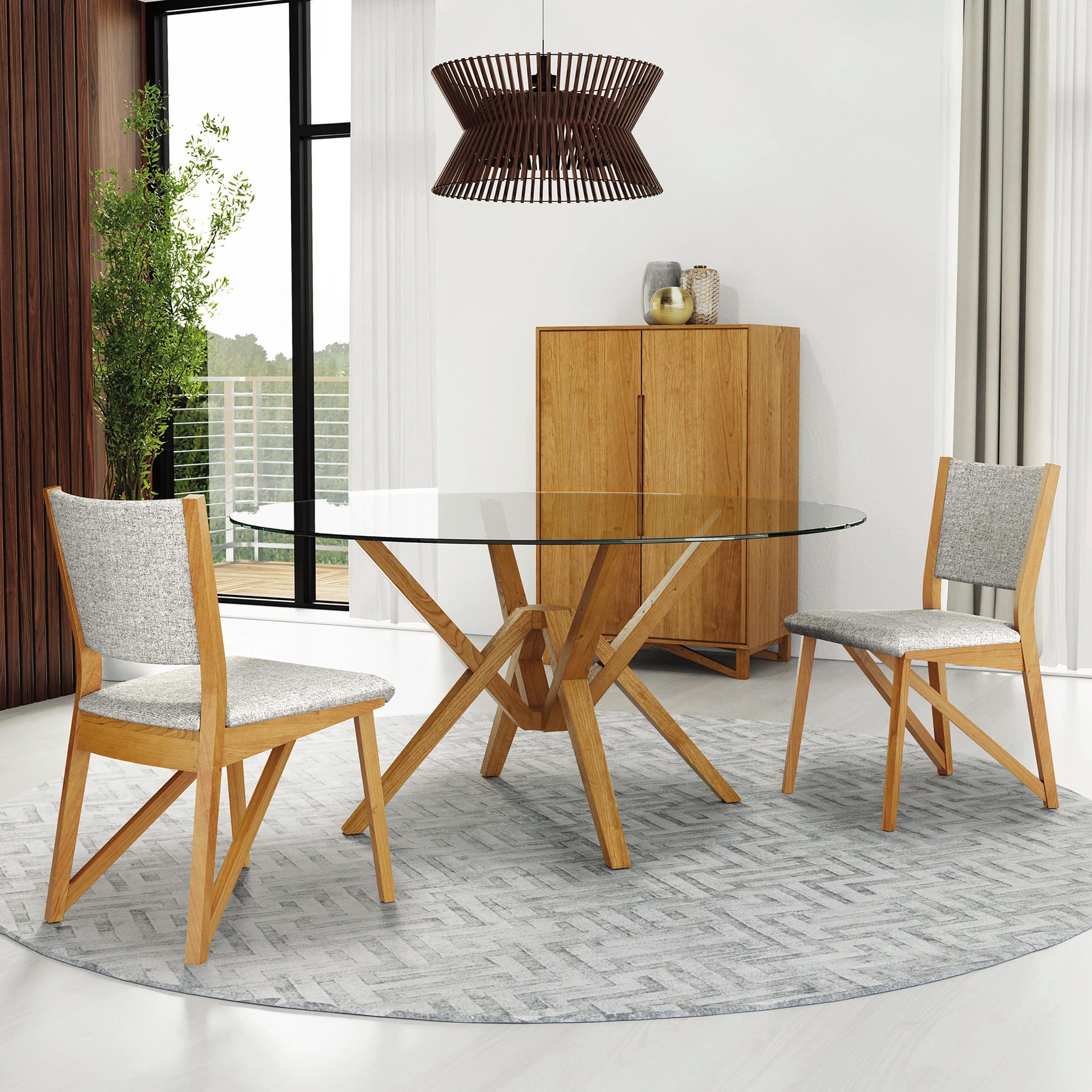 A contemporary dining room features a glass-topped table with a geometric wooden base, surrounded by two Exeter Chairs from Copeland Furniture. A wooden cabinet stands in the corner, and a woven pendant light hangs above. A round rug lies under the table, while a window reveals lush greenery outside.