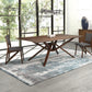 A modern dining room showcases the Exeter Solid Top Dining Table by Copeland Furniture, featuring a wooden design with a geometric base. The space includes two matching chairs with dark upholstered seats, large windows, a contemporary light fixture, a sideboard, and a gray and white patterned rug on a light floor.