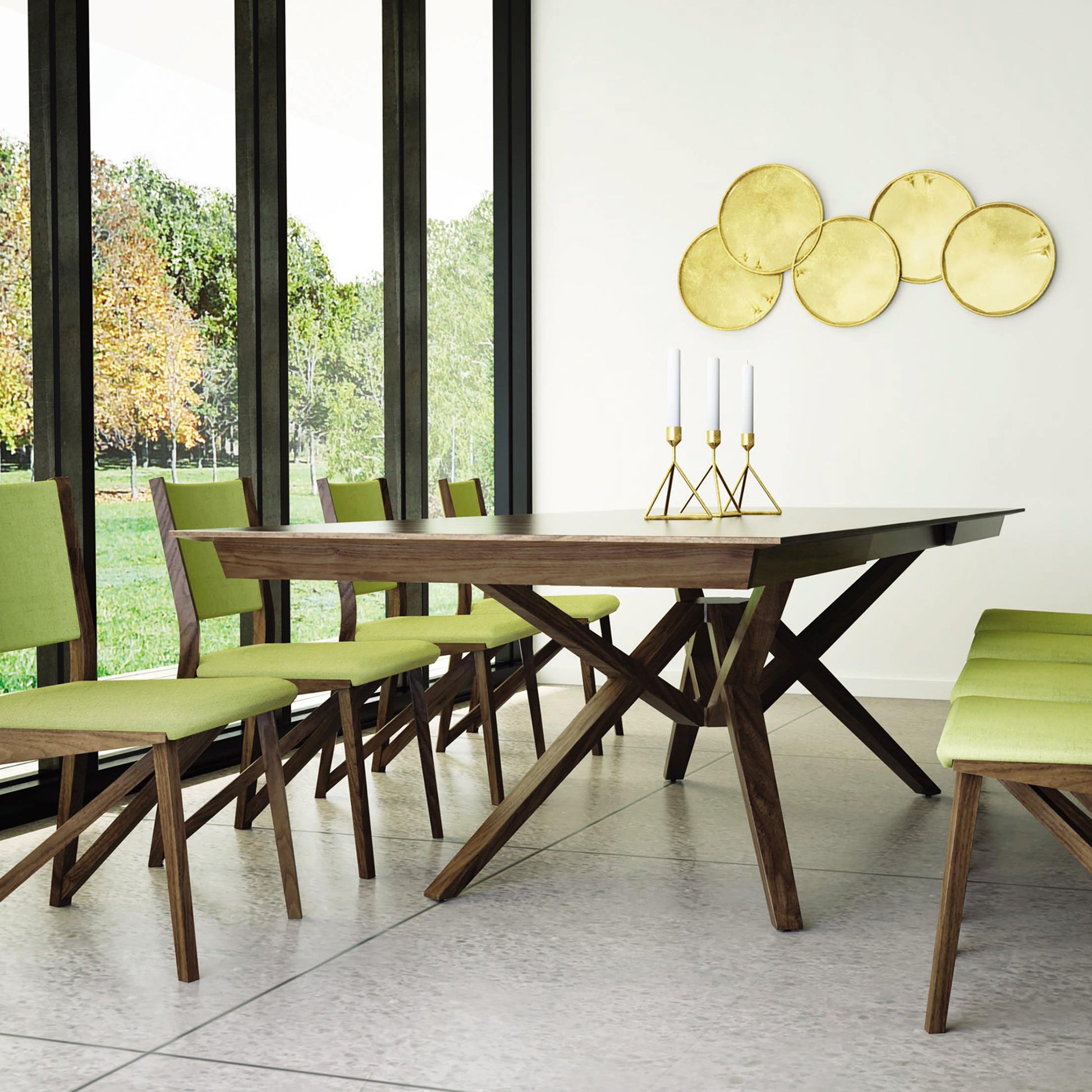 A modern dining room featuring the Exeter Extension Dining Table by Copeland Furniture, made from sustainably harvested hardwood. The wooden table is complemented by green-cushioned wooden chairs. The room boasts large floor-to-ceiling windows, a tiled floor, and gold circular wall decorations with candleholders holding white candles on the table.