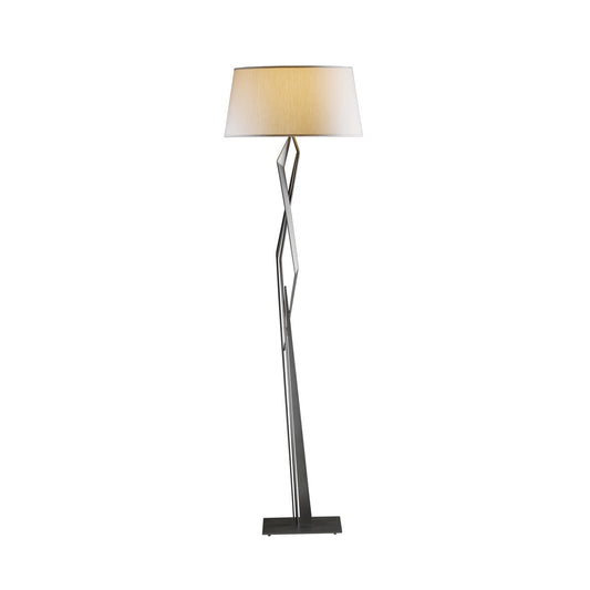 A Hubbardton Forge Facet Floor Lamp with a unique geometric, twisted metal base supporting a large, round, beige lampshade. The lamp stands upright with a simple rectangular base.