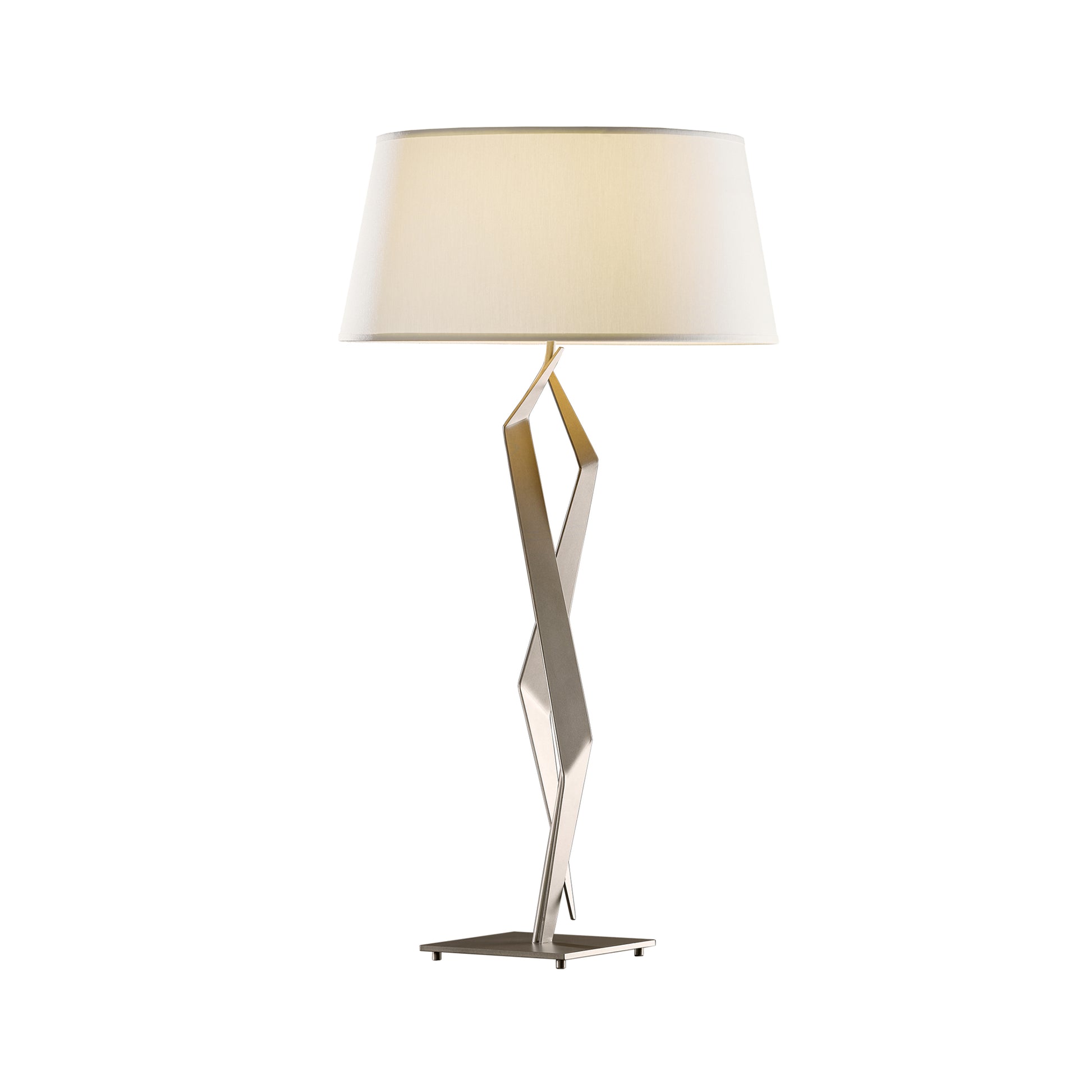 A modern Facet Table Lamp with a zigzag metallic base and a large, round, white lampshade, handcrafted by Hubbardton Forge, isolated on a white background.