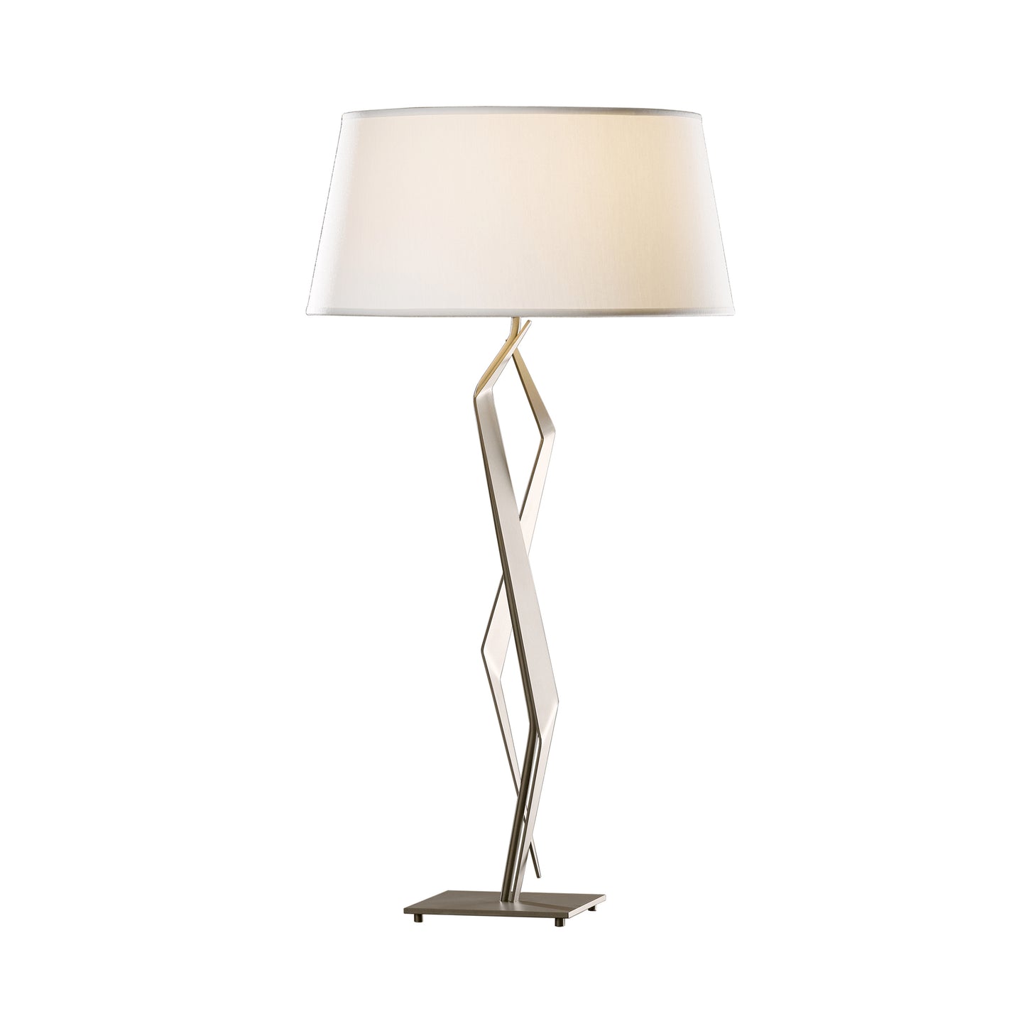 A modern Facet Table Lamp featuring a unique zigzagging metallic base and a large, round, white lampshade, crafted by Hubbardton Forge, standing against a plain white background.