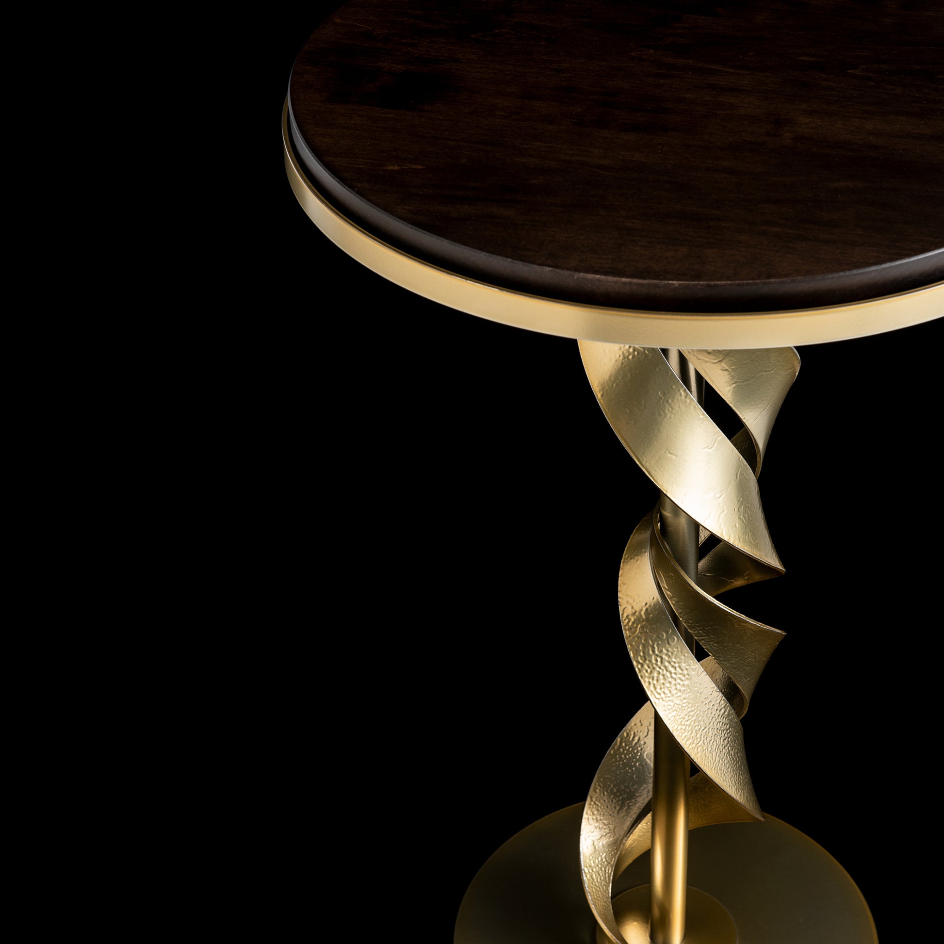 A close-up of the Folio Wood Top Accent Table by Hubbardton Forge showcases its dark Vermont Maple wood top paired with a striking gold, twisted metal stem adorned in a modern brass finish. The black background enhances the contrast between the rich wood tones and the gleaming metallic elements.