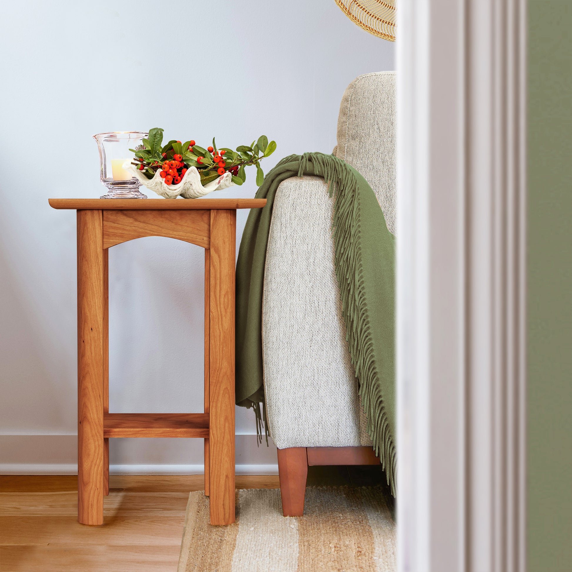 A Heartwood Shaker End Table by Vermont Furniture Designs, crafted from solid wood, is positioned beside a cream-colored sofa decorated with a green throw. Its eco-friendly oil finish shines, drawing attention to a decorative bowl filled with red peppers and leafy greens. The scene is bordered by a wall and doorway on the right.