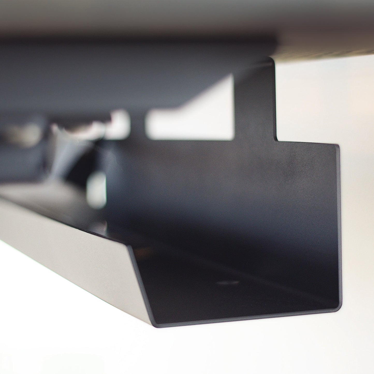 A close-up view of a black metal bracket featuring a rectangular design, mounted under the Invigo Walnut Sit-Stand Desk by Copeland Furniture. The bracket is likely used for organizational or support purposes, showcasing a minimalist design and smooth finish.