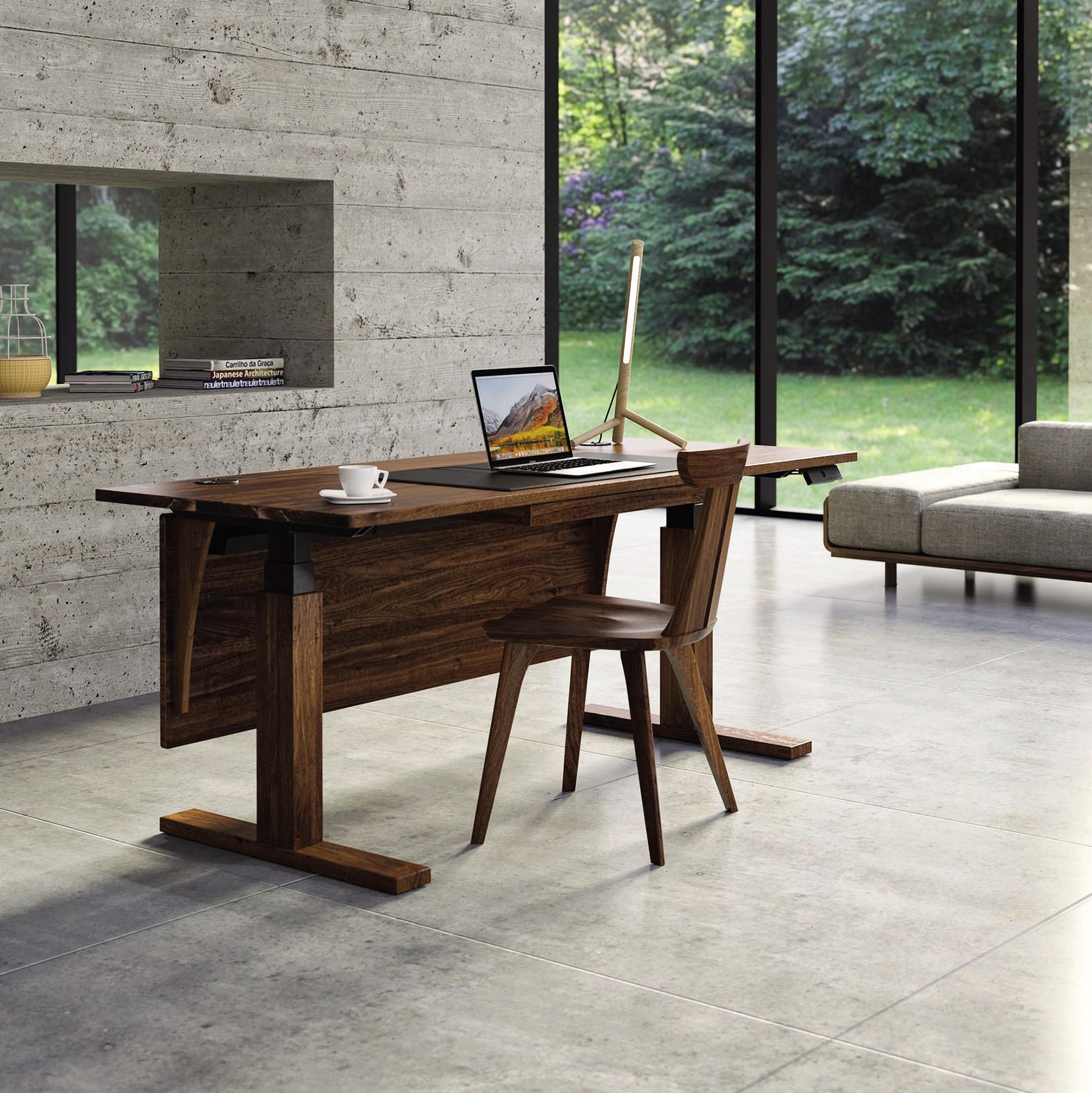 A modern office space features the Invigo Walnut Sit-Stand Desk by Copeland Furniture with programmable height options, equipped with a laptop, desktop monitor, and coffee cup. A wooden chair is positioned at the desk. Large windows provide a view of lush greenery outside, and a light-colored sofa sits to the right.