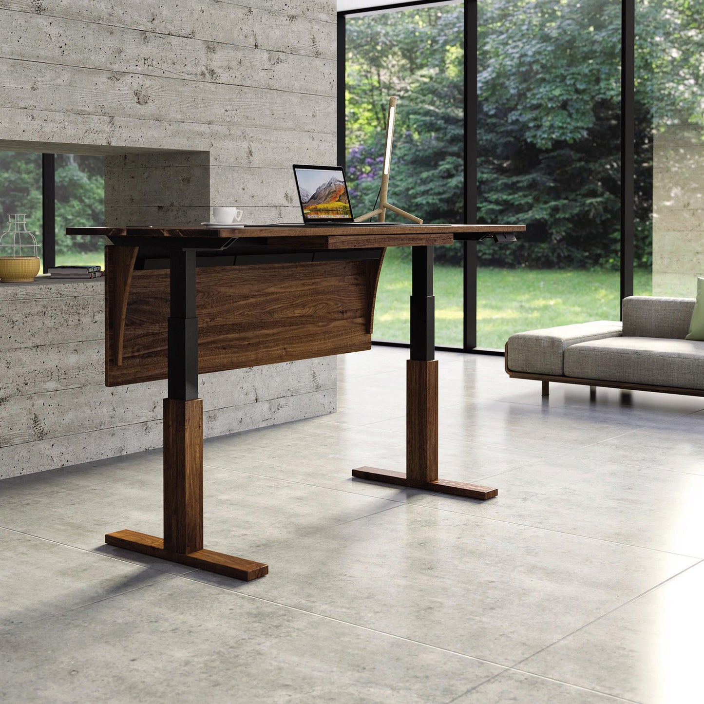 A modern room features the Invigo Walnut Sit-Stand Desk by Copeland Furniture, showcasing solid walnut and black metal legs on a gray tiled floor. The desk, equipped with programmable height options, supports a laptop and a desk lamp. In the background, there is a large window revealing a green garden, while a gray couch is visible on the right.