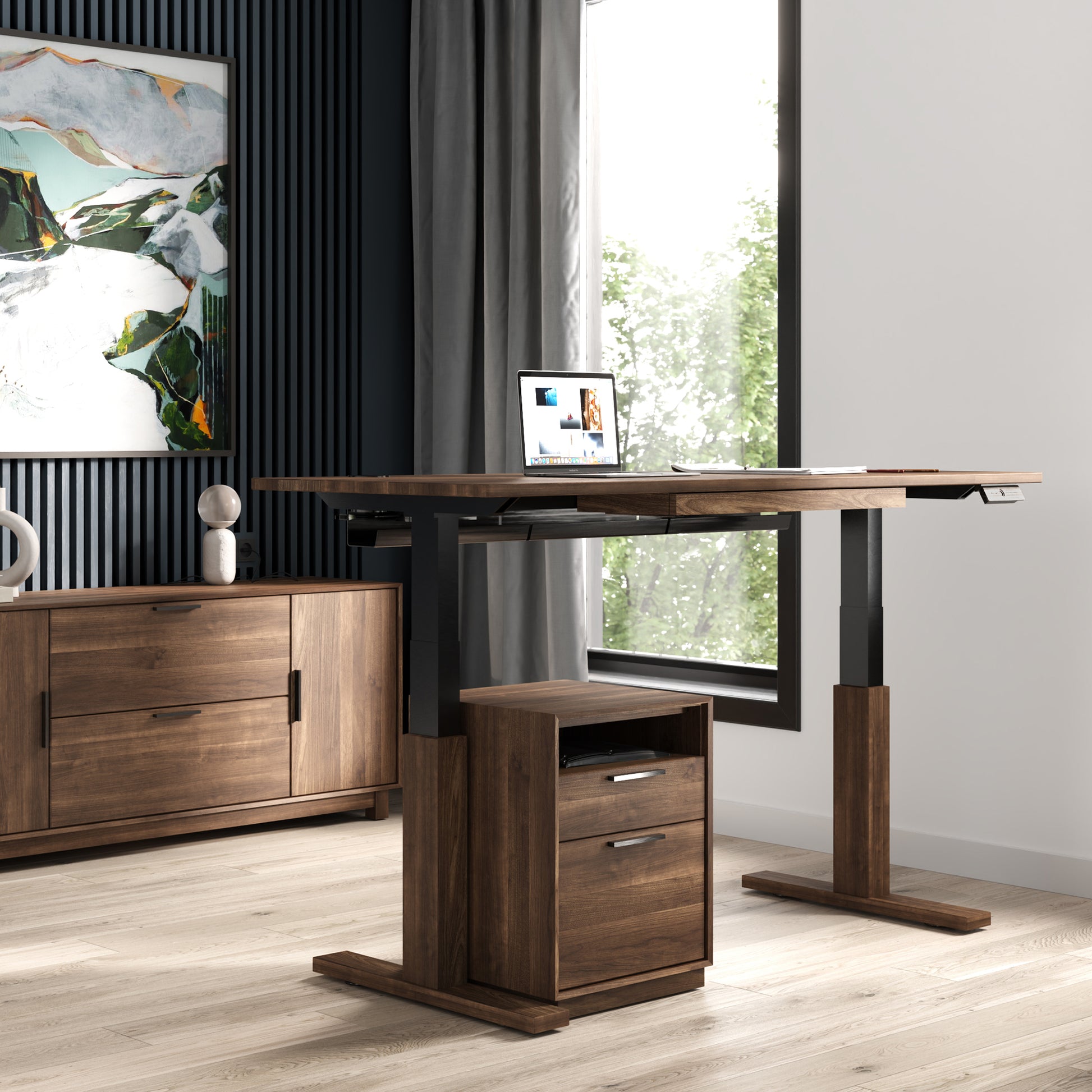 A home office setup featuring the Invigo Walnut Sit-Stand Desk by Copeland Furniture, showcasing a laptop and some paperwork. The room has a large window with greenery outside, a wooden filing cabinet under the desk, and a matching wooden sideboard with abstract artwork above it.