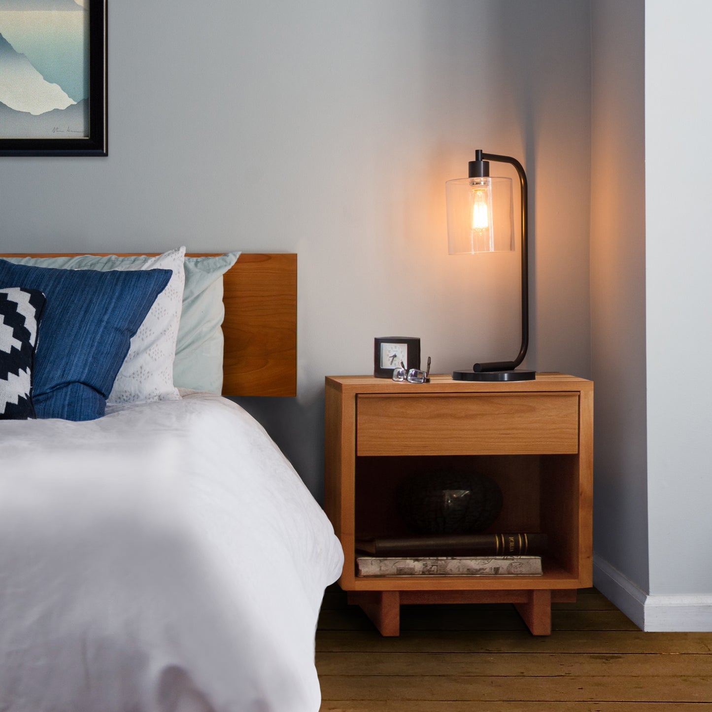 A cozy bedroom corner featuring a Vermont Furniture Designs Kipling 1-Drawer Enclosed Shelf Nightstand with a lamp, photo frame, and decorative items, next to a bed with blue and white bedding.