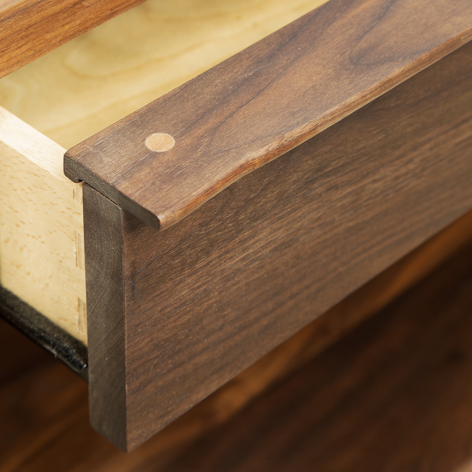 Close-up of an open wooden drawer showcasing dovetail joinery in the Kipling 1-Drawer Enclosed Shelf Wide Nightstand by Vermont Furniture Designs. The front panel is crafted from rich walnut with visible grain, while the interior features lighter natural maple. A round wooden plug adorns the top corner of the drawer front.