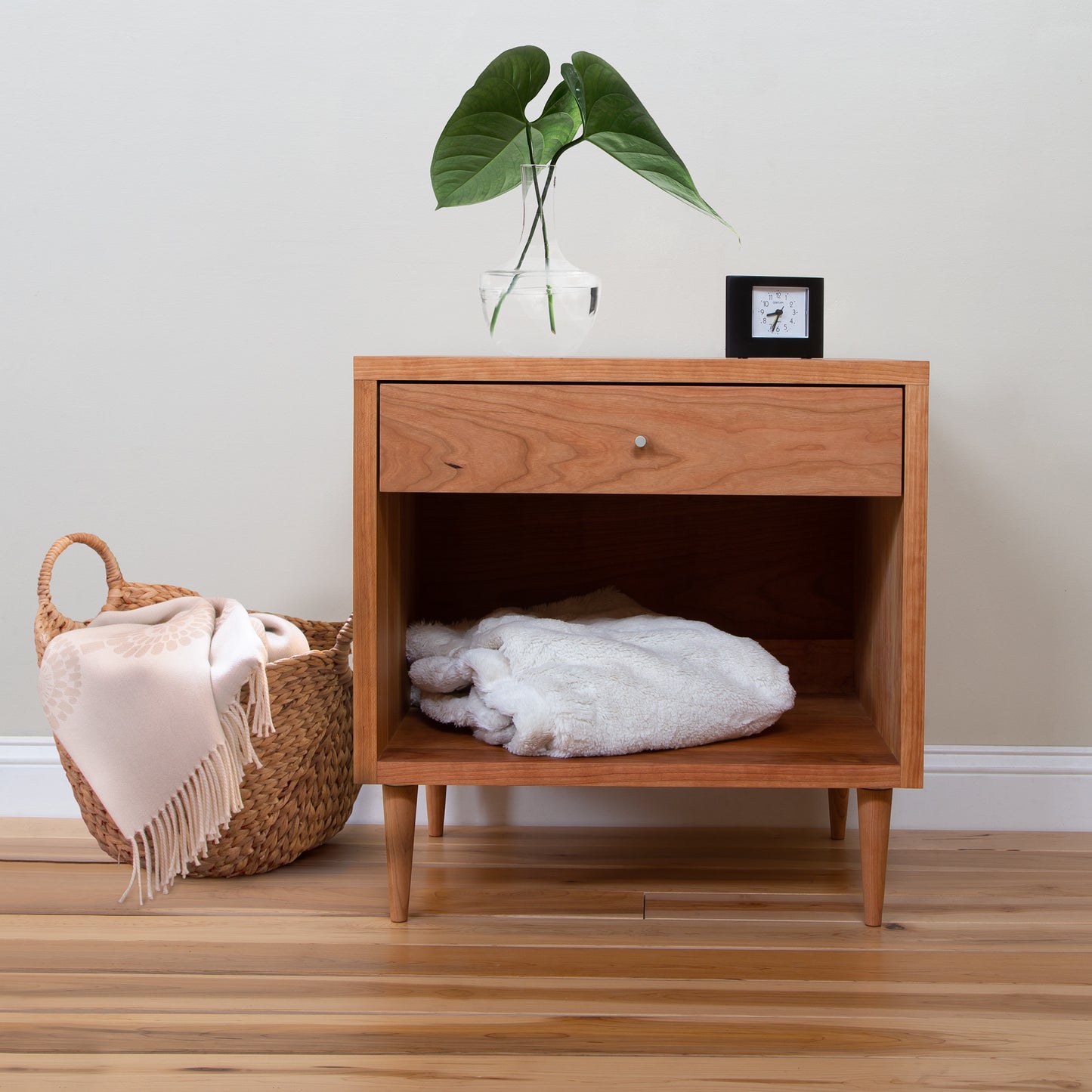 A Larssen 1-Drawer Wide Nightstand following a Mid-Century Modern Design, with an open drawer containing folded towels, next to a wicker basket with a draped blanket, under a hanging plant, against a neutral.
