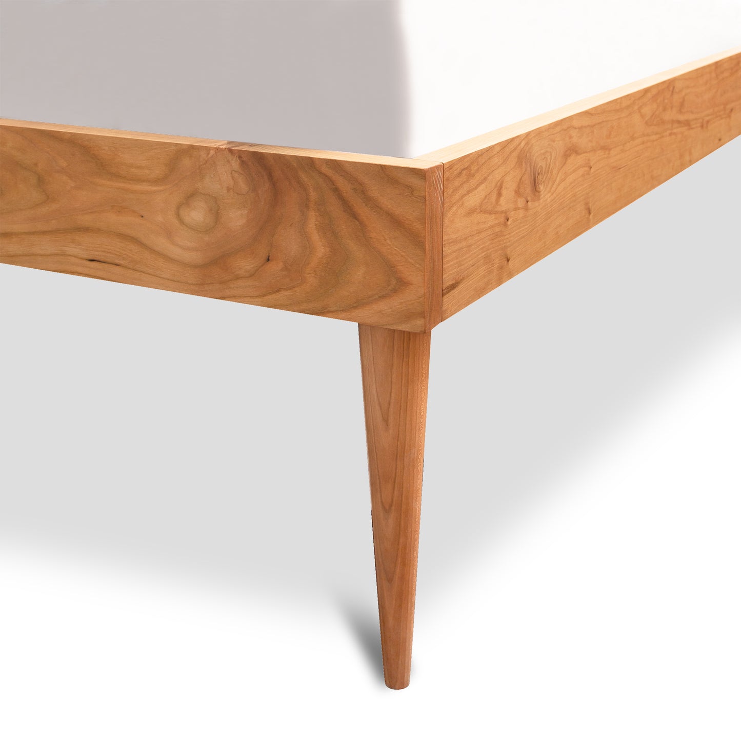 Close-up of the corner of a solid wood bed frame in a modern bedroom. The Larssen Bed - Queen - Ready to Ship by Vermont Furniture Designs features a natural cherry wood finish with visible wood grain patterns and sleek, tapered legs. A white mattress peeks over the top edge of the frame. Plain, light-colored background. American made furniture from Vermont Woods Studios.