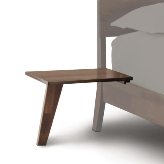 The Linn Attached Nightstand by Copeland Furniture, crafted from walnut and inspired by mid-century modern design, seamlessly integrates with the bed frame. Featuring a single angled leg for support and a smooth, flat surface, this ready-to-ship piece is shown alongside a partial view of the bed frame and mattress with bedding.