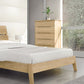 A minimalist bedroom featuring a wooden bed frame, white bedding, and the Linn 5-Drawer Wide Chest by Copeland Furniture made from upcycled North American hardwood. A small plant in a metallic pot sits on top of the chest. A large window on the right allows natural light to fill the room, highlighting its mid-century modern design.