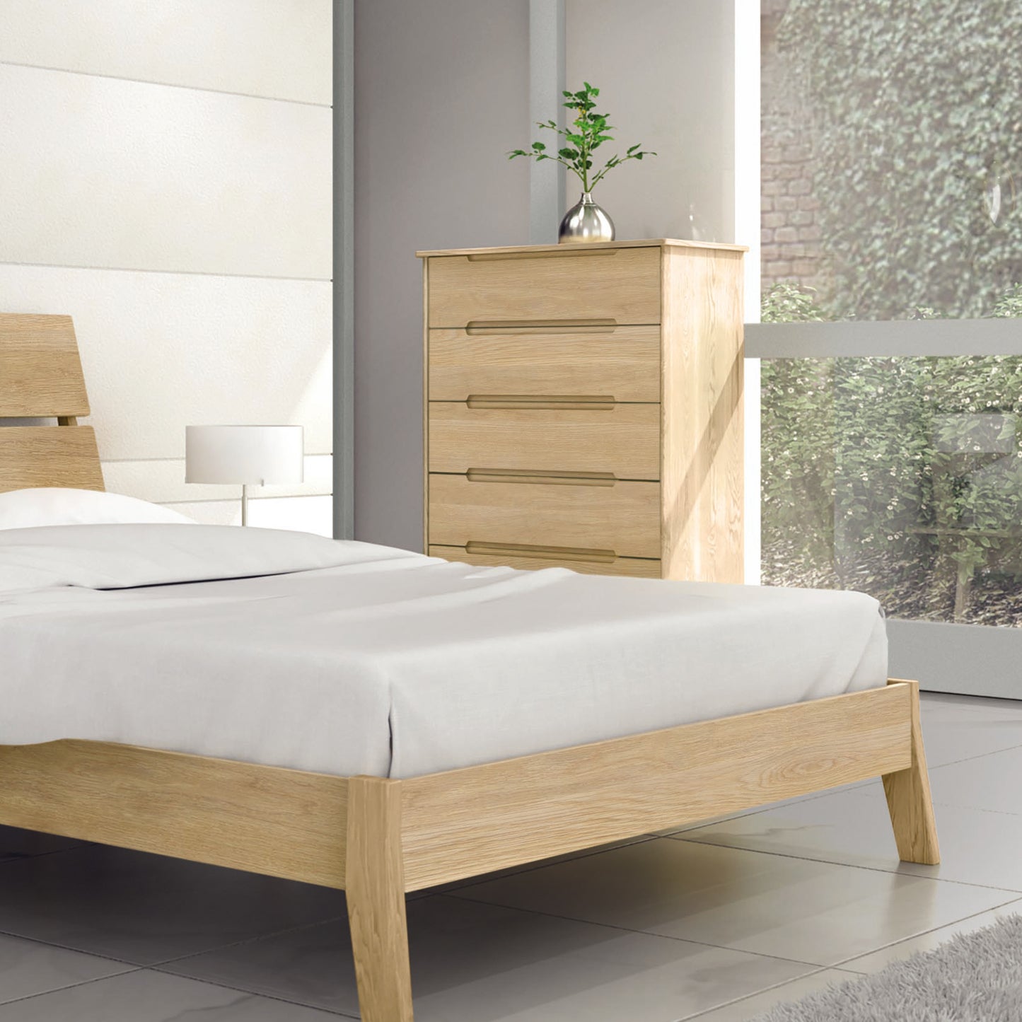 A minimalist bedroom featuring a wooden bed frame, white bedding, and the Linn 5-Drawer Wide Chest by Copeland Furniture made from upcycled North American hardwood. A small plant in a metallic pot sits on top of the chest. A large window on the right allows natural light to fill the room, highlighting its mid-century modern design.