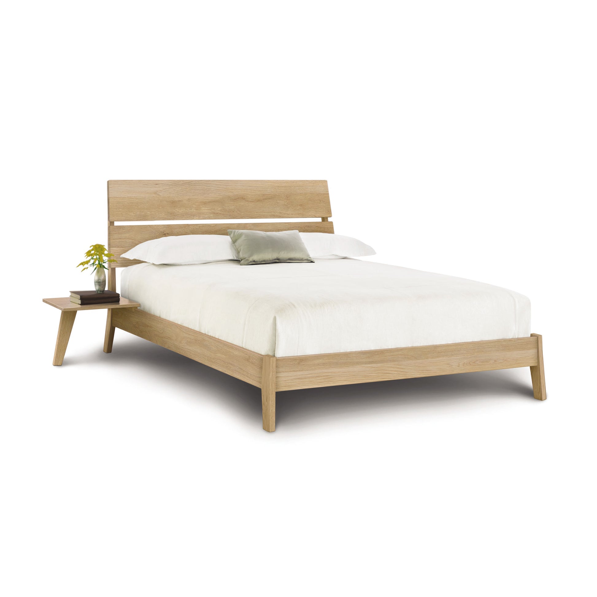 A Linn Oak Platform Bed frame with a headboard, dressed in white bedding, accompanied by a small bedside table with a green plant, isolated against a white background. (Copeland Furniture)