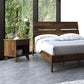 A bedroom featuring a wooden bed frame made from sustainable solid wood and the Linn 1-Drawer Enclosed Shelf Nightstand by Copeland Furniture. The bed is dressed with white sheets and pillows, while the nightstand holds a lamp and offers an additional small shelf underneath for storage. Large windows reveal greenery outside, contrasting with the sleek concrete floor.