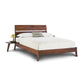 The Linn Walnut Platform Bed by Copeland Furniture is a stunning piece crafted from upcycled wood, featuring a matching headboard and adorned with crisp white linens. This designer bed includes a built-in side table on the left, which holds a vase with vibrant yellow flowers. A single green pillow adds a pop of color to the white blanket, and the elevated bed frame proudly carries Greenguard certification for sustainability.