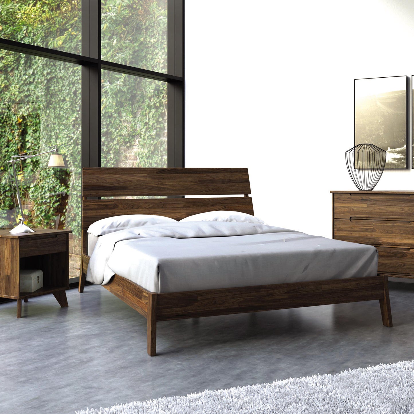 A modern bedroom features the Linn Walnut Platform Bed by Copeland Furniture, crafted from upcycled wood with a gray comforter. The space includes a matching wooden nightstand topped with a metallic lamp, complemented by a white floor rug and wooden dresser. Designer touches include a decorative wire sculpture and a framed picture. Large windows provide views of the greenery outside.