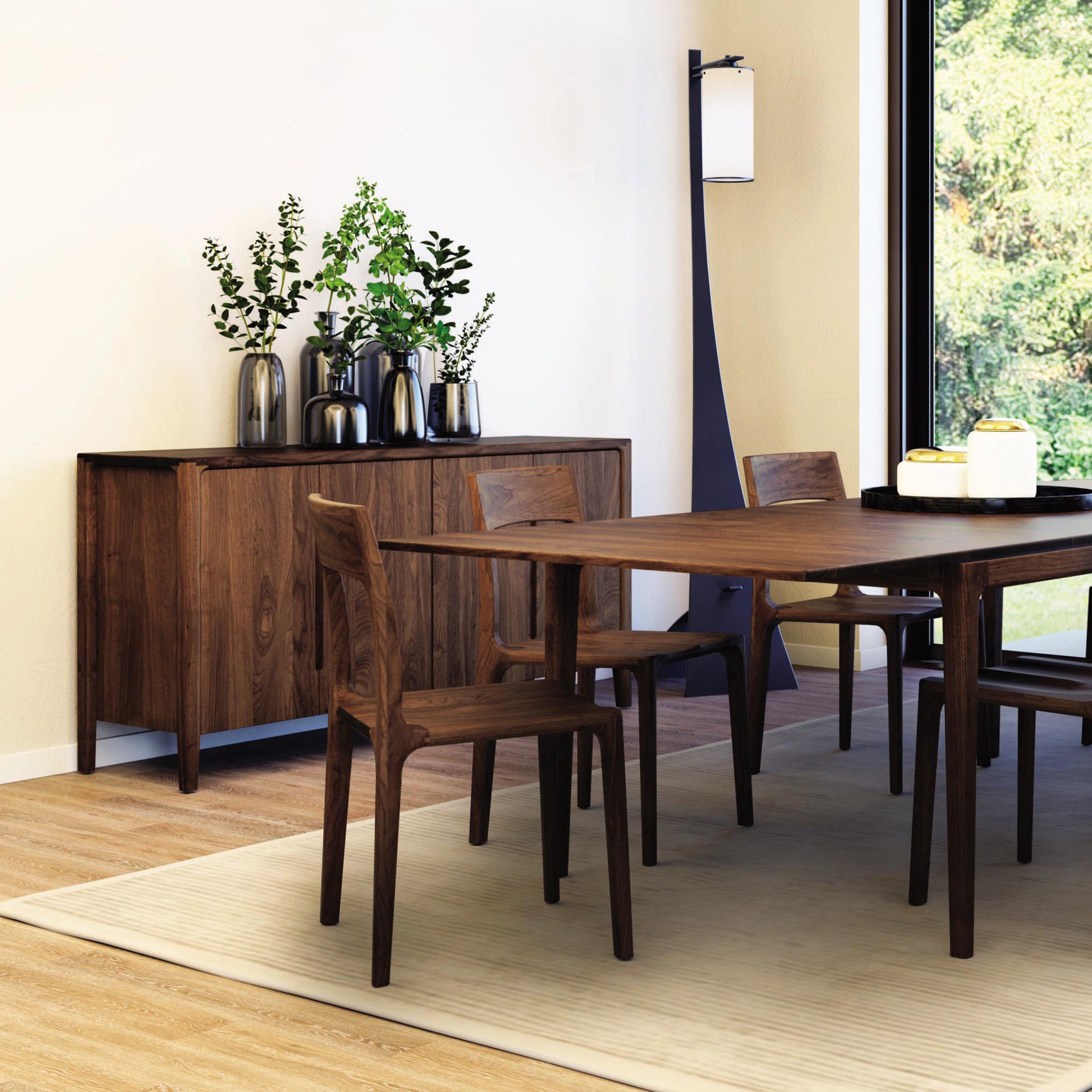 A dining room features a wooden table with four matching chairs on a beige rug. In the background, a Lisse Buffet by Copeland Furniture displays decorative black vases and green plants. Nearby, a tall floor lamp stands next to sliding glass doors that open up to an outdoor view, enhancing the modern design.