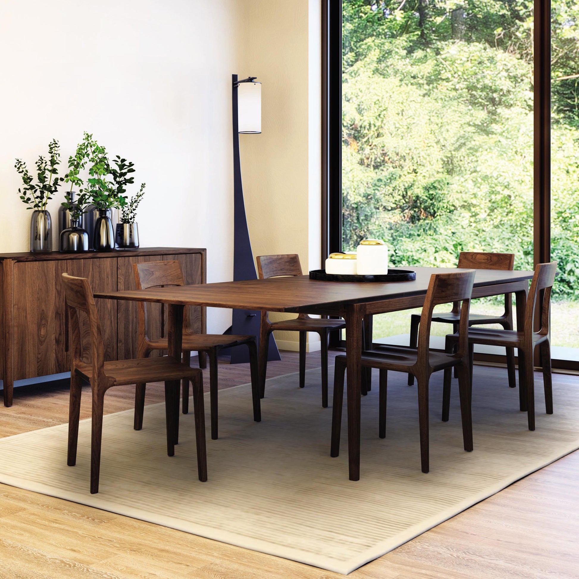 A dining room features the Lisse Extension Dining Table by Copeland Furniture, accompanied by six matching chairs, all set on a beige rug. A sideboard adorned with decorative vases and greenery stands against the wall, while large windows in the background reveal a view of lush green foliage outside.