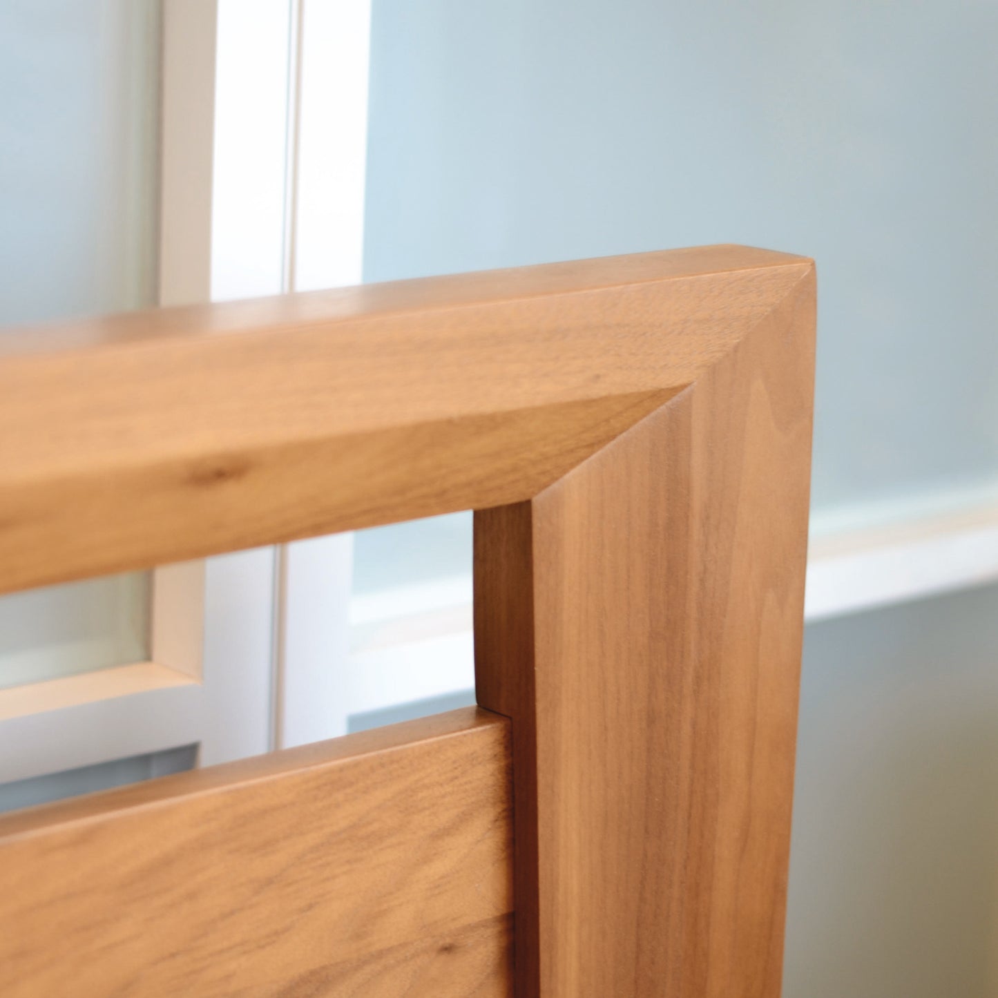 Close-up view of the corner of the Mansfield Cherry Platform Bed, handmade in Vermont by Copeland Furniture from solid cherry wood, with a smooth, light brown finish against a backdrop of frosted and clear glass panels.