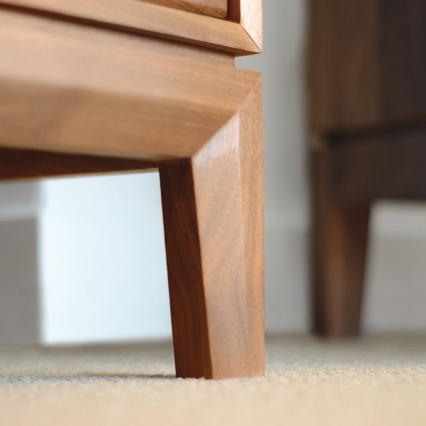 Close-up of the Mansfield 1-Drawer Enclosed Shelf Nightstand by Copeland Furniture, showcasing its wooden leg made from smooth, light brown organic solid wood. The leg exhibits clean, straight lines and sharp angles, positioned on a beige carpet with a blurred background that reveals another piece of eco-friendly furniture in a similar style.