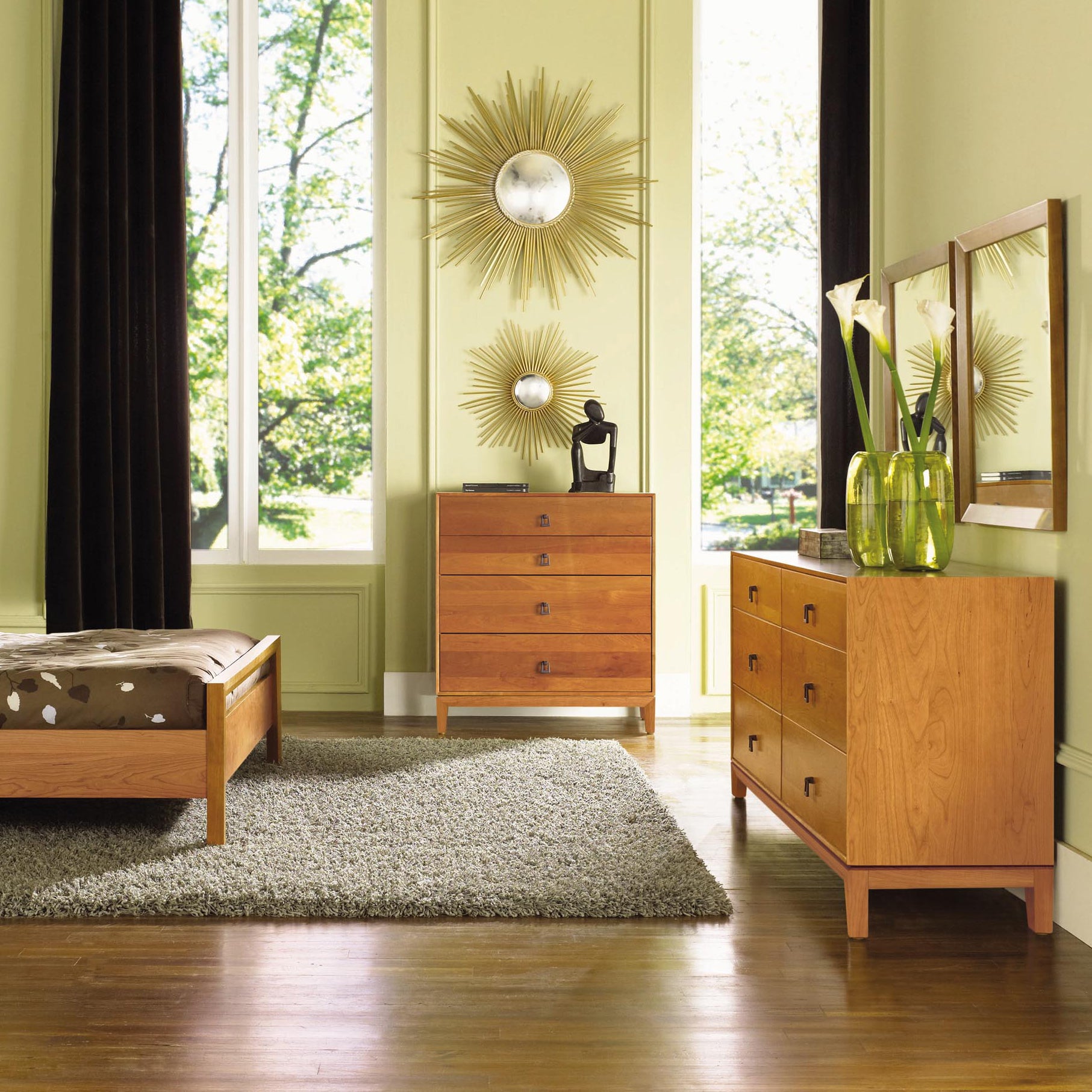 A bedroom with light green walls, a large window with dark curtains, and solid wood furniture including a bed, a Copeland Furniture Mansfield 6-Drawer Dresser, and a chest of drawers. The room also features a gray rug, a sunburst mirror, and a vase with white flowers on the dresser.