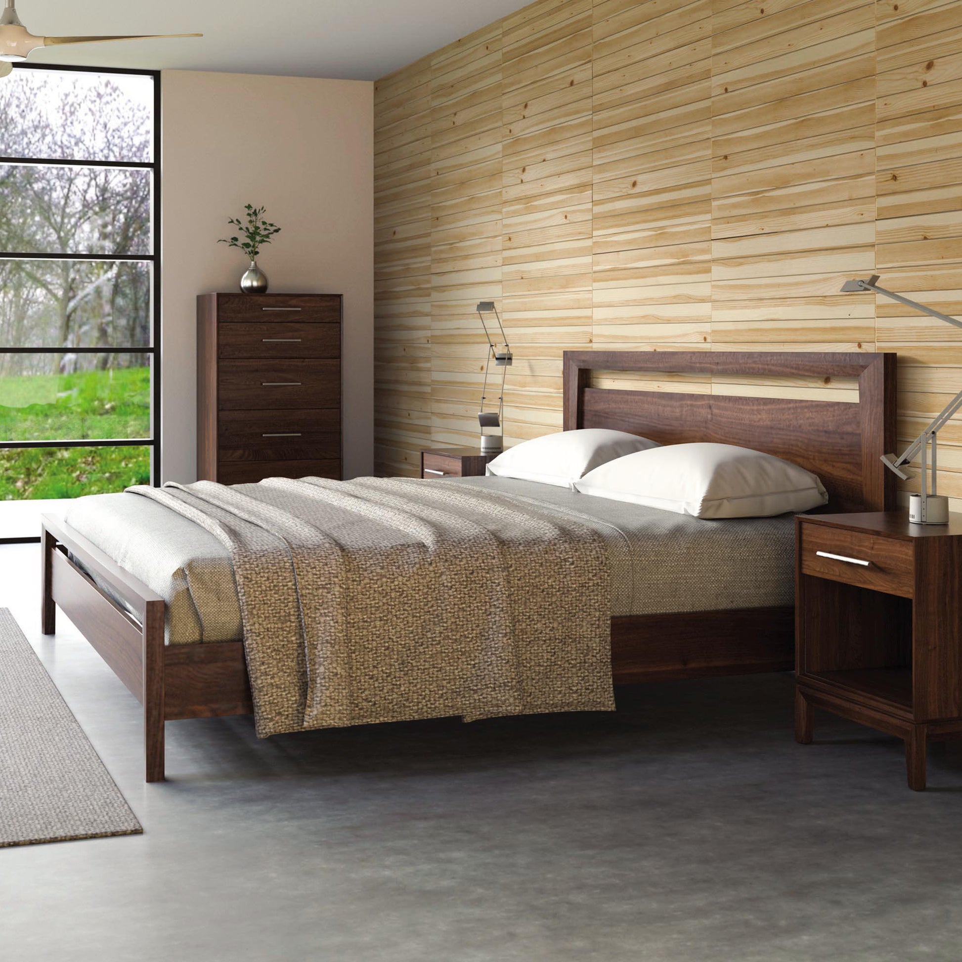 A bedroom features the Copeland Furniture Mansfield Walnut Platform Bed with beige bedding and a textured throw blanket. Two matching wooden nightstands with lamps, a dresser beside the bed, and a large window revealing greenery outside complete the Arts & Crafts style room, with wood-paneled walls behind the bed.