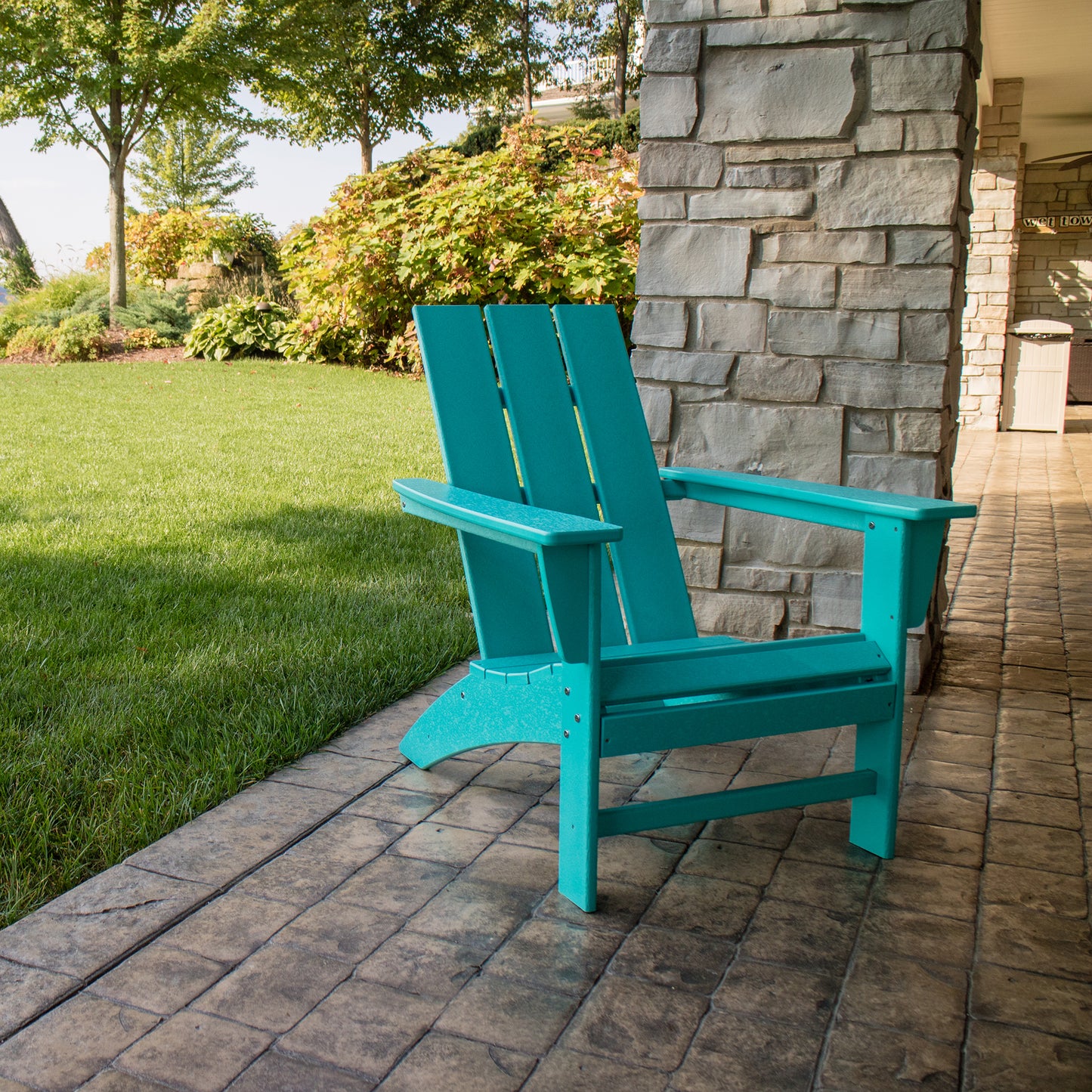 A turquoise POLYWOOD Modern Adirondack Chair is positioned on a stone patio, next to a vibrant green lawn. Made from durable POLYWOOD lumber, the patio is framed by a stone wall, with trees and shrubs visible in the background beneath a clear sky.