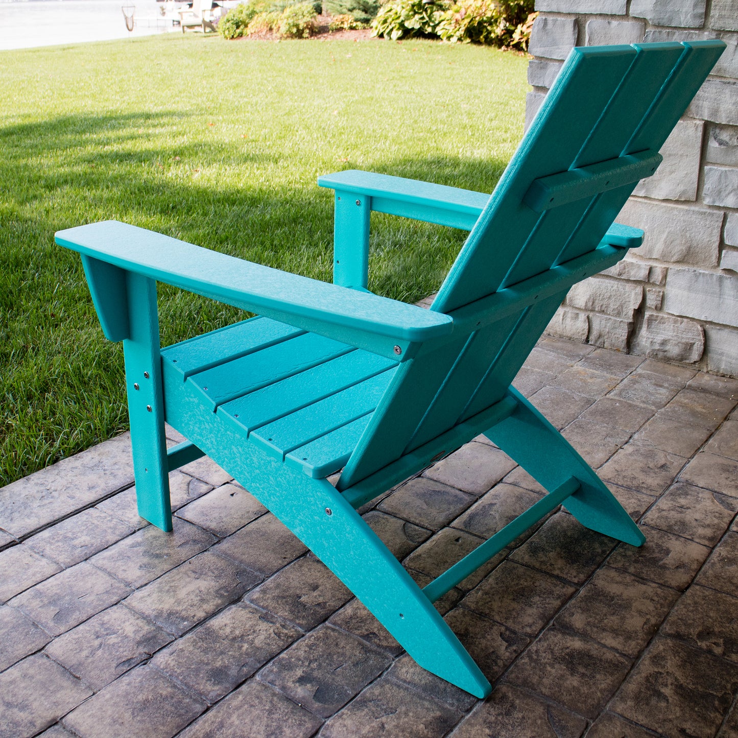 A POLYWOOD Modern Adirondack Chair in weather-resistant turquoise is placed on a stone patio, crafted from durable POLYWOOD material. It offers a view of a well-kept lawn next to a stone wall adorned with lush greenery, creating a tranquil and serene outdoor ambiance.