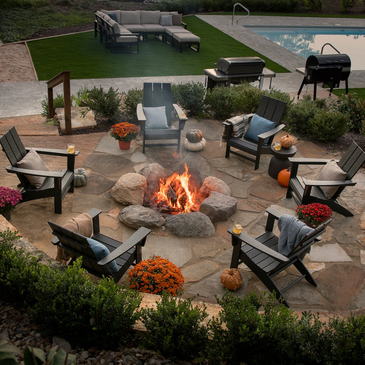 In a backyard scene, a stone patio highlights a weather-resistant fire pit surrounded by six POLYWOOD Modern Adirondack Chairs. The setting is further enhanced with pumpkins and potted flowers, while a pool and additional outdoor furniture complete the serene atmosphere.