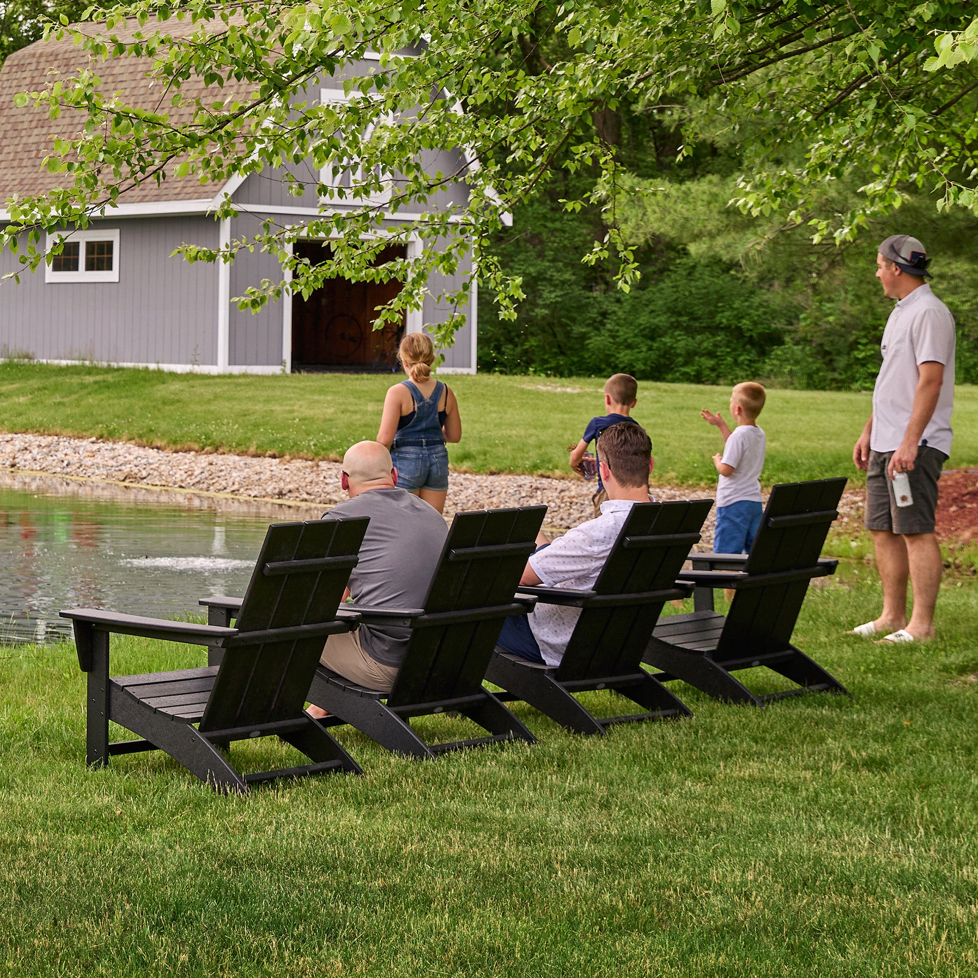 A group of people is gathered outdoors by a pond. Three individuals are seated on POLYWOOD Modern Adirondack Chairs, made from weather-resistant POLYWOOD lumber, facing the water. Meanwhile, two adults and three children stand closer to the shore. In the background stands a gray barn surrounded by trees and grass.