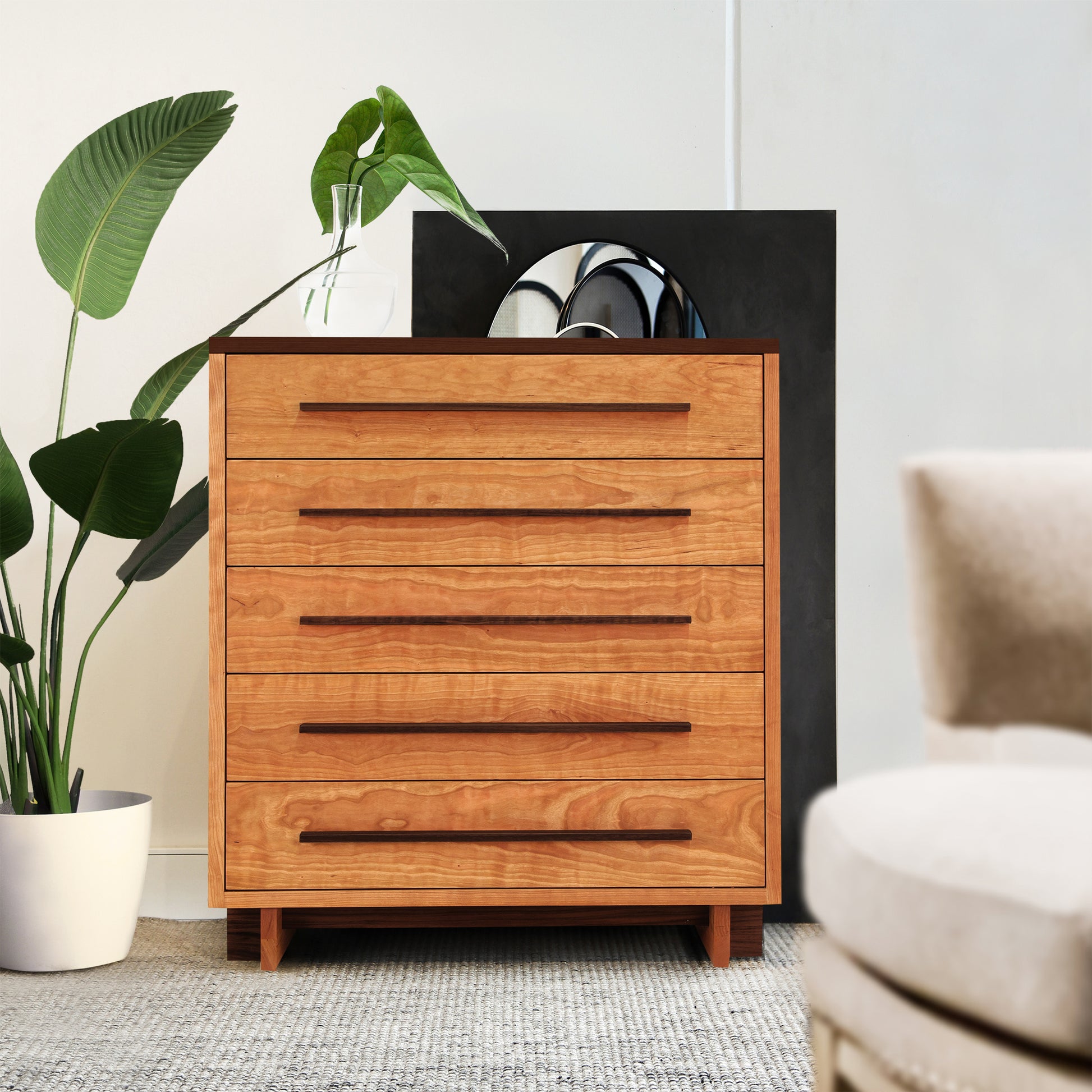 A Vermont Furniture Designs Modern American 5-Drawer Chest with curved handles in a minimalist room features an eco-friendly oil finish. A potted plant and a black decorative element are visible behind the dresser. Part of a beige chair appears in