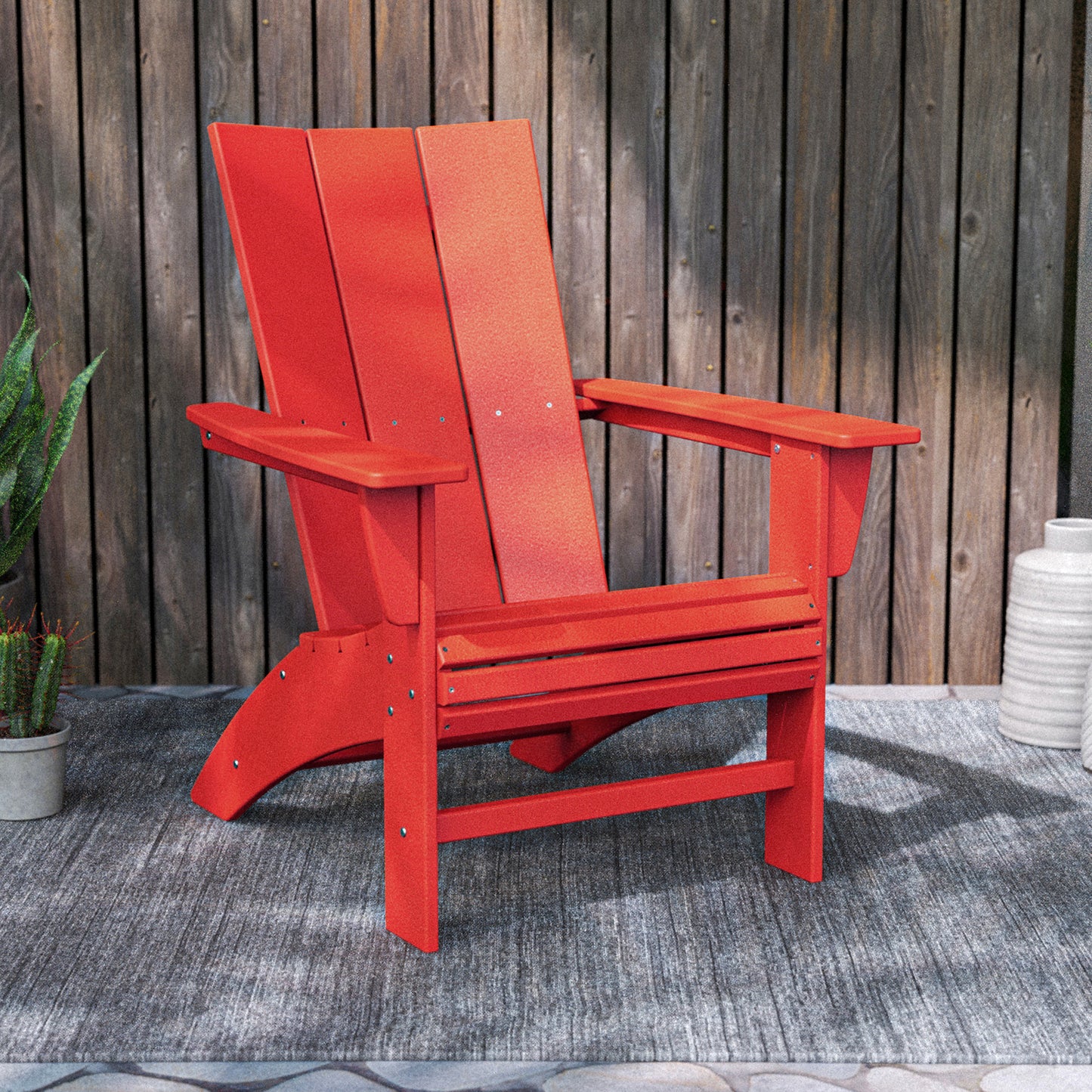 A vibrant red POLYWOOD Modern Curveback Adirondack Chair, made from robust POLYWOOD lumber, rests on a gray outdoor rug in a welcoming outdoor area. Situated in front of a wooden fence and accompanied by a potted plant and white ceramic pot, it enhances the backyard's charm.