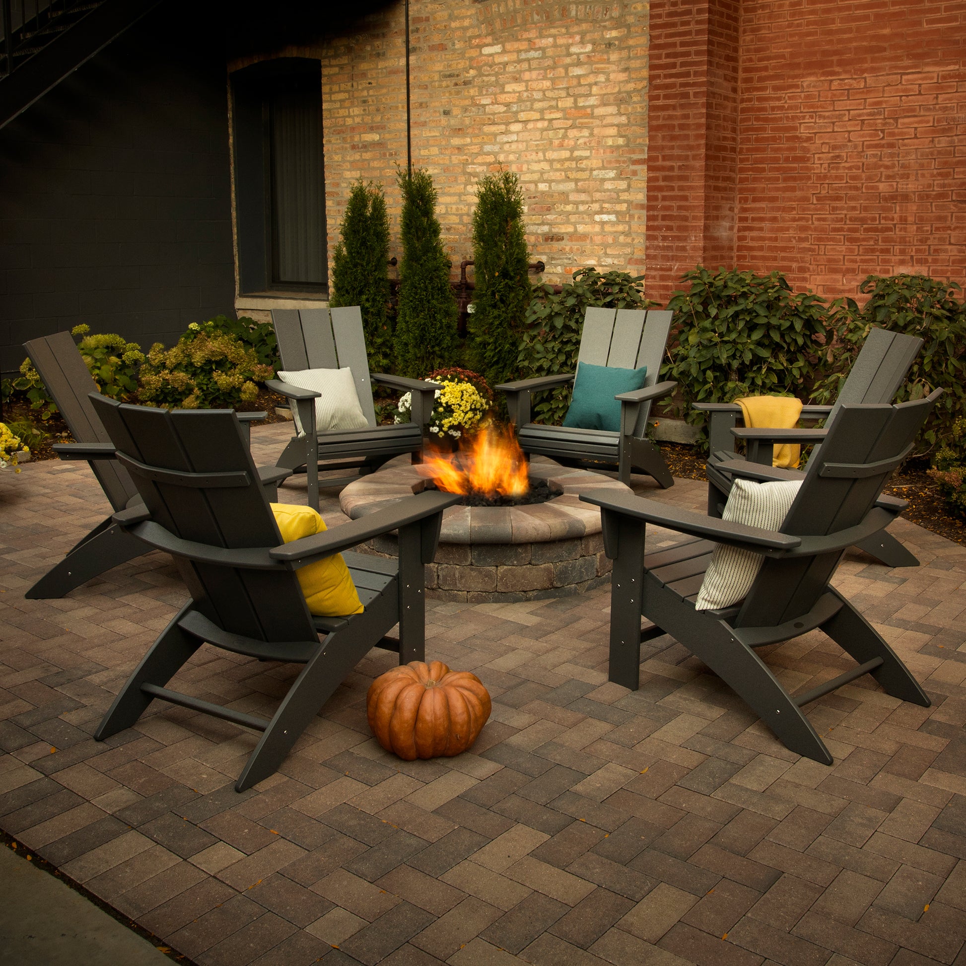 Six Modern Curveback Adirondack Chairs by POLYWOOD, crafted from durable POLYWOOD® lumber, encircle a stone fire pit with a warm, flickering flame. The chairs are enhanced with cushions, and a pumpkin is stationed in front. Brick walls and potted plants offer the ideal backdrop for this inviting outdoor setting.