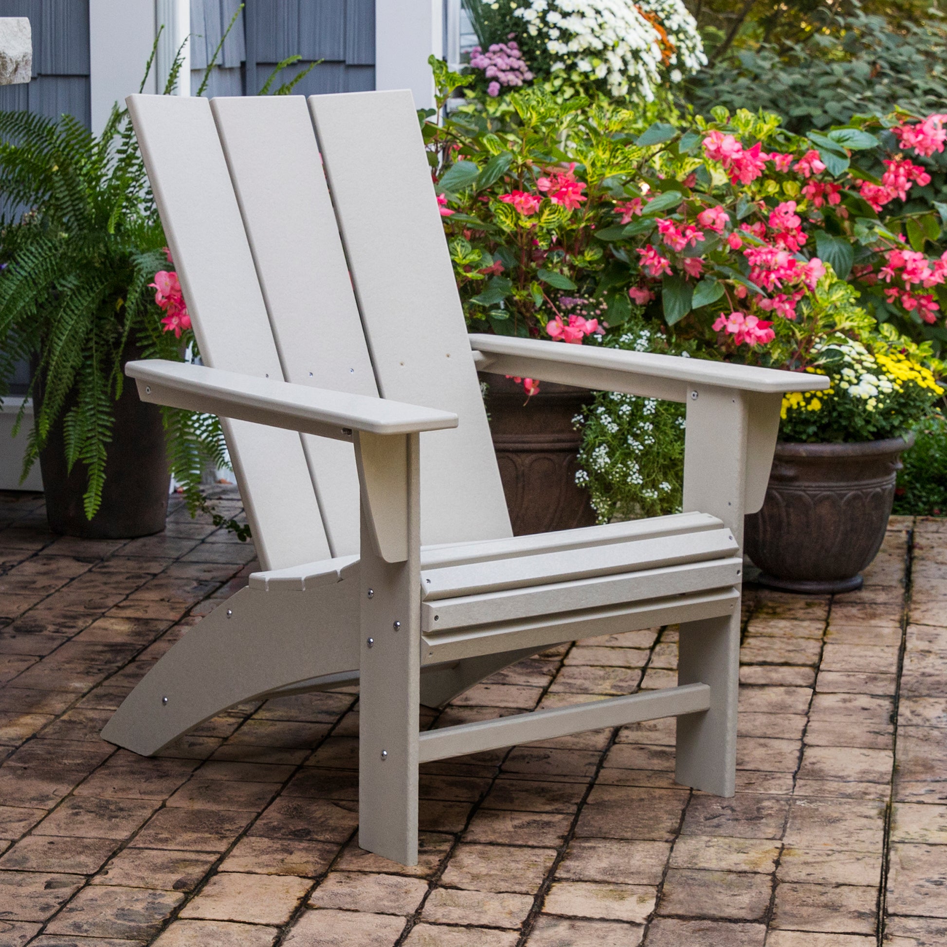 A POLYWOOD Modern Curveback Adirondack Chair, crafted from durable POLYWOOD lumber, sits on a brick patio, enveloped by an inviting outdoor setting. Potted plants, adorned with pink and white flowers and lush green ferns, thrive nearby against a backdrop of a gray building wall.