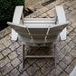 Top view of a POLYWOOD Modern Curveback Adirondack Chair expertly crafted from POLYWOOD® lumber, positioned on a textured stone-paved patio. The chair is situated close to a garden area adorned with lush green shrubs. Enhancing the outdoor setting, a rectangular grate is embedded within the pavement.