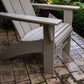 The POLYWOOD Modern Curveback Adirondack Chair, crafted from durable POLYWOOD® lumber, sits elegantly on a stone patio surrounded by lush green bushes and vibrant yellow flowers. The outdoor setting is enhanced by the chair's angled backrest slats and wide armrests.