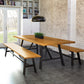 A modern dining room features a wooden table with two matching Modern Farmhouse Benches from Copeland Furniture. Above the table hangs a black dome-shaped light fixture. The brick walls and large windows give a rustic, airy feel. A potted plant from Copeland Furniture sits at the center of the table.