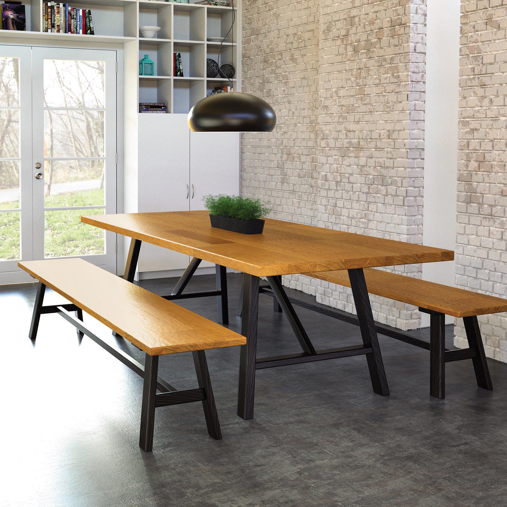 A modern dining room features a wooden table with two matching Modern Farmhouse Benches from Copeland Furniture. Above the table hangs a black dome-shaped light fixture. The brick walls and large windows give a rustic, airy feel. A potted plant from Copeland Furniture sits at the center of the table.