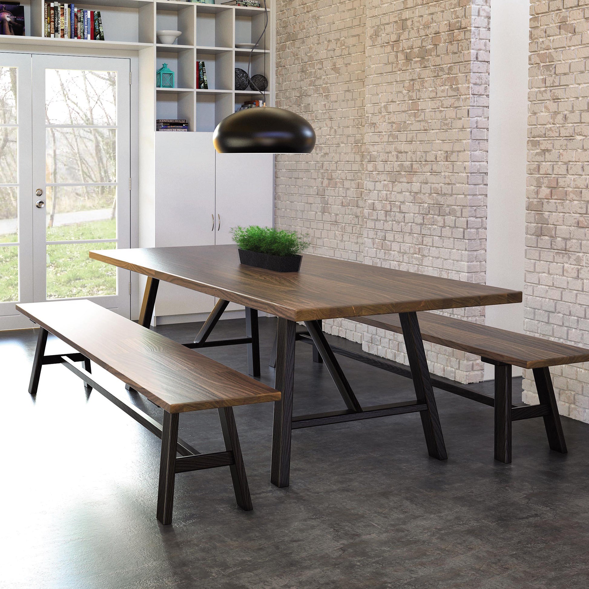 A modern dining area features a wooden table reminiscent of a Modern Farmhouse Trestle Farm Table, flanked by two matching benches. The table rests on a dark floor next to a brick wall with a large window and glass doors in the background. A black pendant light from Copeland Furniture illuminates the space.