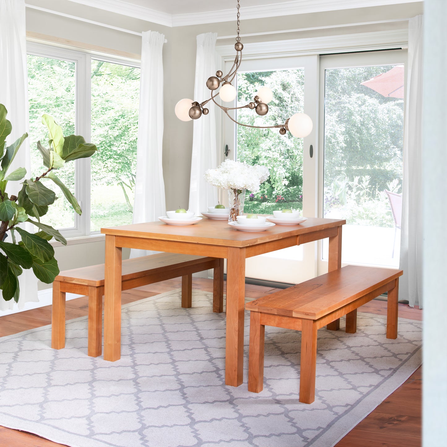 Alt text: Elegant dining room featuring a solid wood table and two matching American Mission Benches by Lyndon Furniture. Modern chandelier with round bulbs above, large windows, sliding glass door, white curtains, potted plant on patterned rug for enhanced décor. High quality American made furniture in cherry and walnut wood.