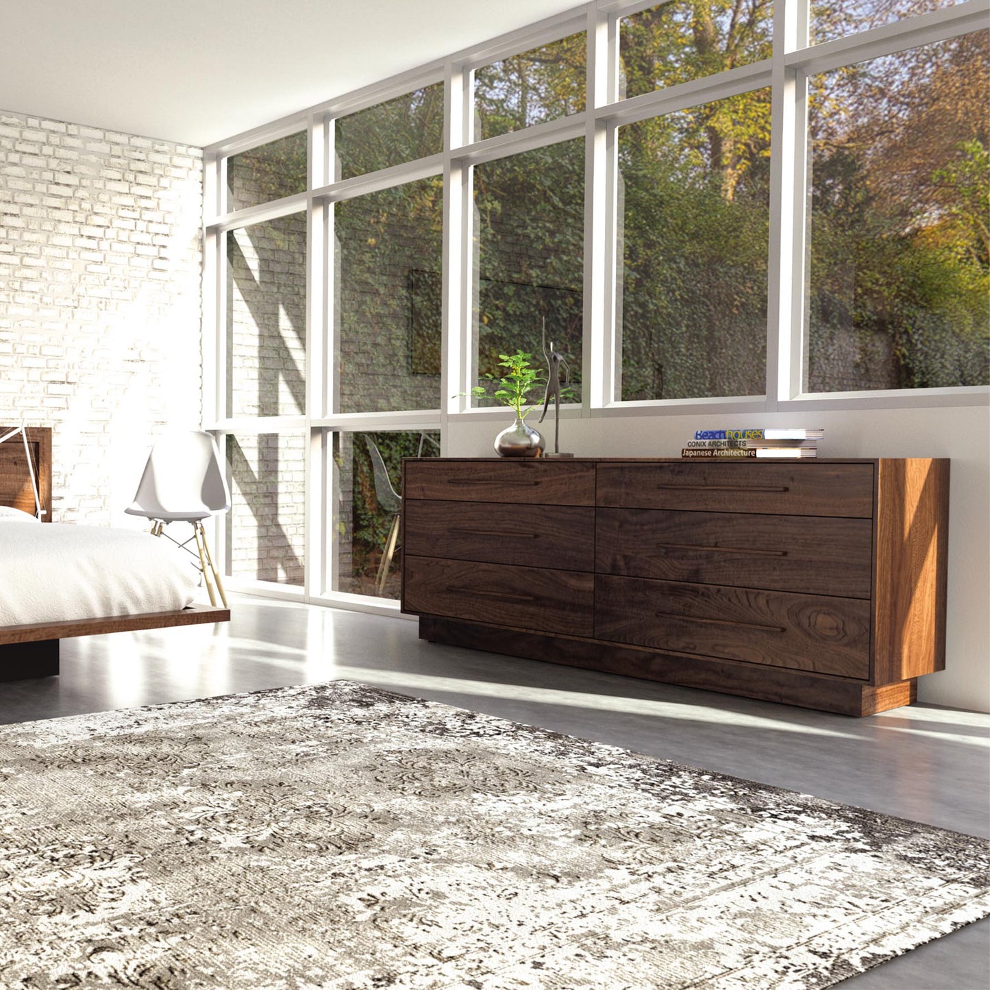 A modern bedroom featuring a large window with a view of trees, a white bed, a white chair, and a large patterned rug. The room has a bright and airy ambiance, complemented by the sustainably harvested natural woods of the Copeland Furniture Moduluxe 6-Drawer Dresser - 29" Series with books and a plant on top.