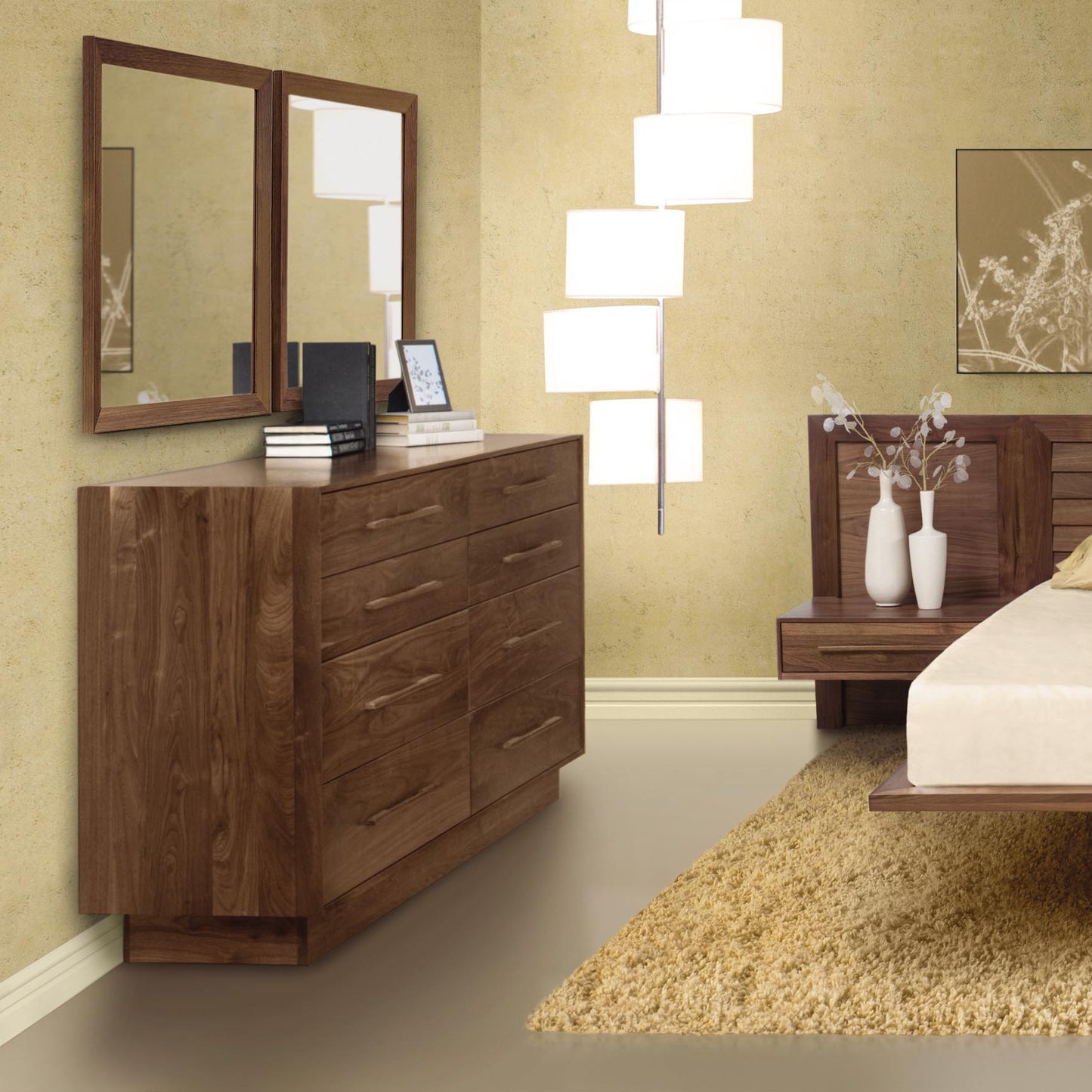 A bedroom with beige walls features a solid wood Copeland Furniture Moduluxe 8-Drawer Dresser - 35" Series from the eco-friendly Moduluxe Bedroom Furniture collection, topped with two framed mirrors. The dresser holds books and a picture frame. A nightstand with a vase of white flowers is beside the bed with white linens, and a multi-light floor lamp stands nearby.